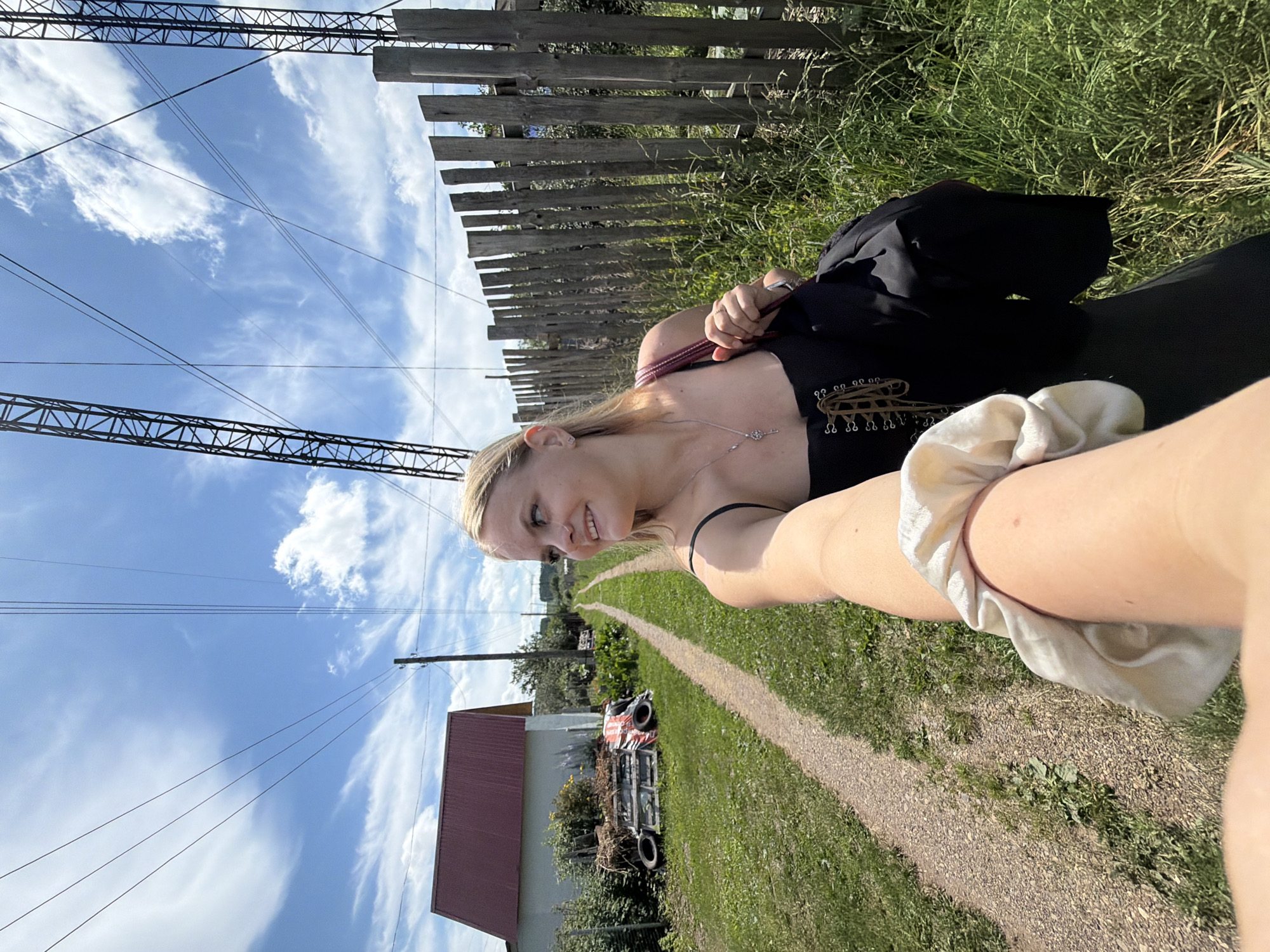 In the city of Chaikovsky, Russia, a young woman with blonde hair is captured in a selfie outdoors. She is wearing a black corset-style top with visible lacing and a silver necklace. The background reveals a rural setting with a dirt path, green grass, and a wooden fence. To the left, a red-roofed building stands, with some discarded items like tires and pallets nearby. Overhead, power lines stretch across a bright blue sky dotted with white clouds. The woman is smiling and looking to her right, holding a dark-colored bag and wearing a scrunchie on her arm.