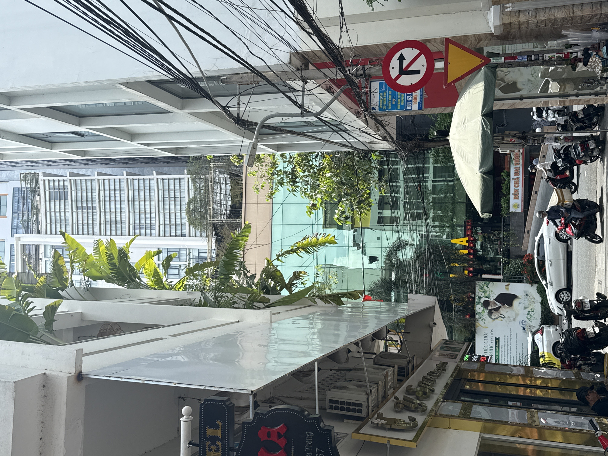 Here's a detailed description of the image:
**Setting:** This is a street scene, likely in the Nha Trang Ward area of Nha Trang City, Vietnam.
**Foreground:**
* A building with a white, layered awning or rooftop is prominent.
* Vegetation is growing on the rooftop structure, including banana trees.
* A sign for "The" is partly visible.
* Displays of merchandise, possibly shoes, are set out on shelves.
**Midground:**
* A street scene with motorcycles parked along the sidewalk is visible.
* A sign for "Bún Chả Hai Thường" (a Vietnamese noodle dish restaurant) is seen, along with a poster depicting a wedding and the text "TIỆC CUỐI PHÚC VẸN TRÒN" in Vietnamese.
* A white car is parked.
* People are present near the storefront.
**Background:**
* Modern buildings, including one with glass facade reflecting the sky.
* Overhead power lines are tangled in the upper part of the frame.
* Traffic signs are visible, including a "No Left Turn" sign and a yellow triangular yield sign.
