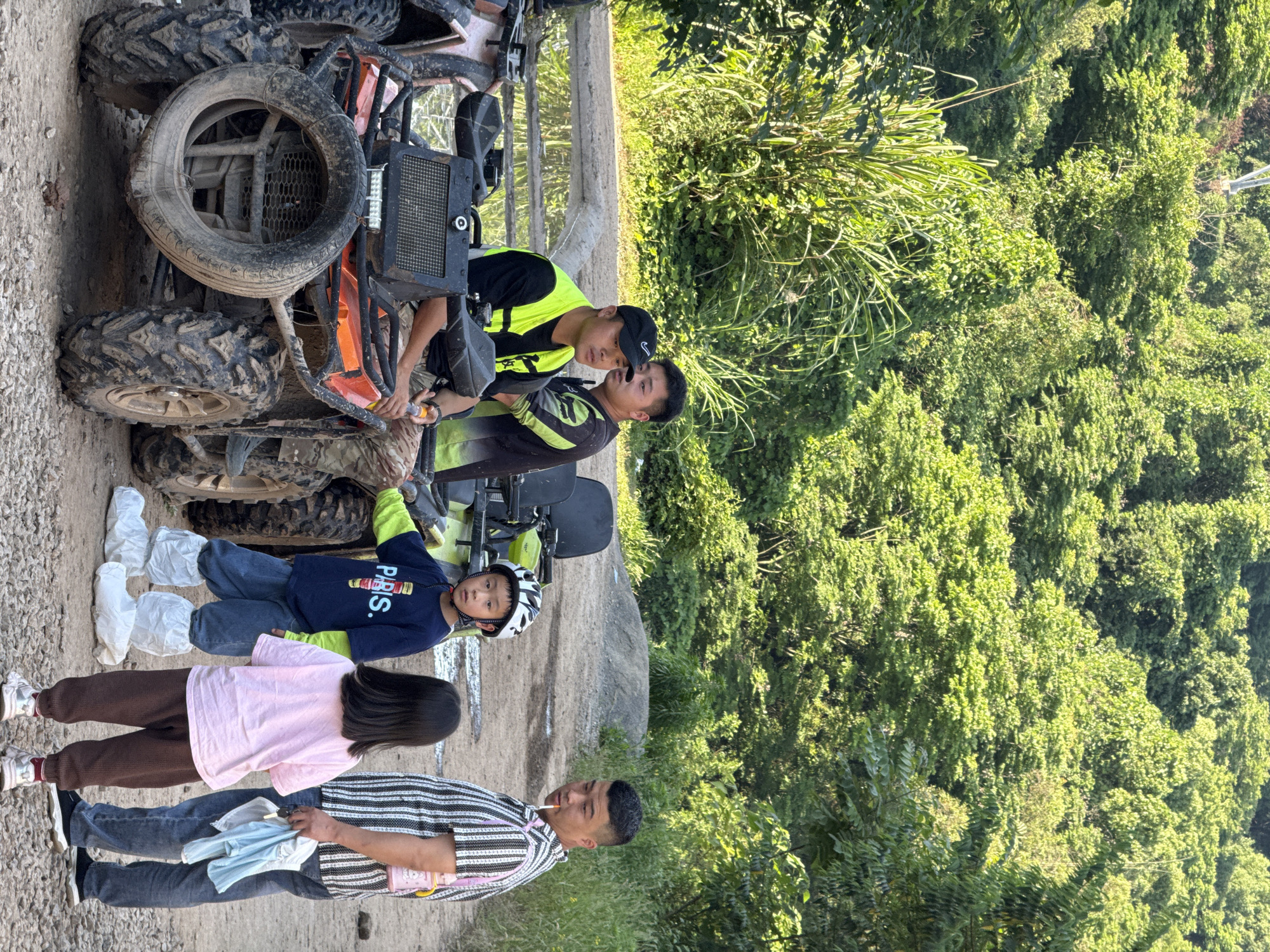 The image shows a group of people standing next to an ATV. There are four individuals: two men, a girl, and a boy. The men are standing near the ATV, which is on a dirt path next to a lush green forest. One man is wearing a safety vest, and the other is wearing a striped shirt. The boy is wearing a helmet and booties. The girl is wearing a pink shirt and brown pants. The location appears to be outdoors, possibly a recreational area. The city is Beibei, China.