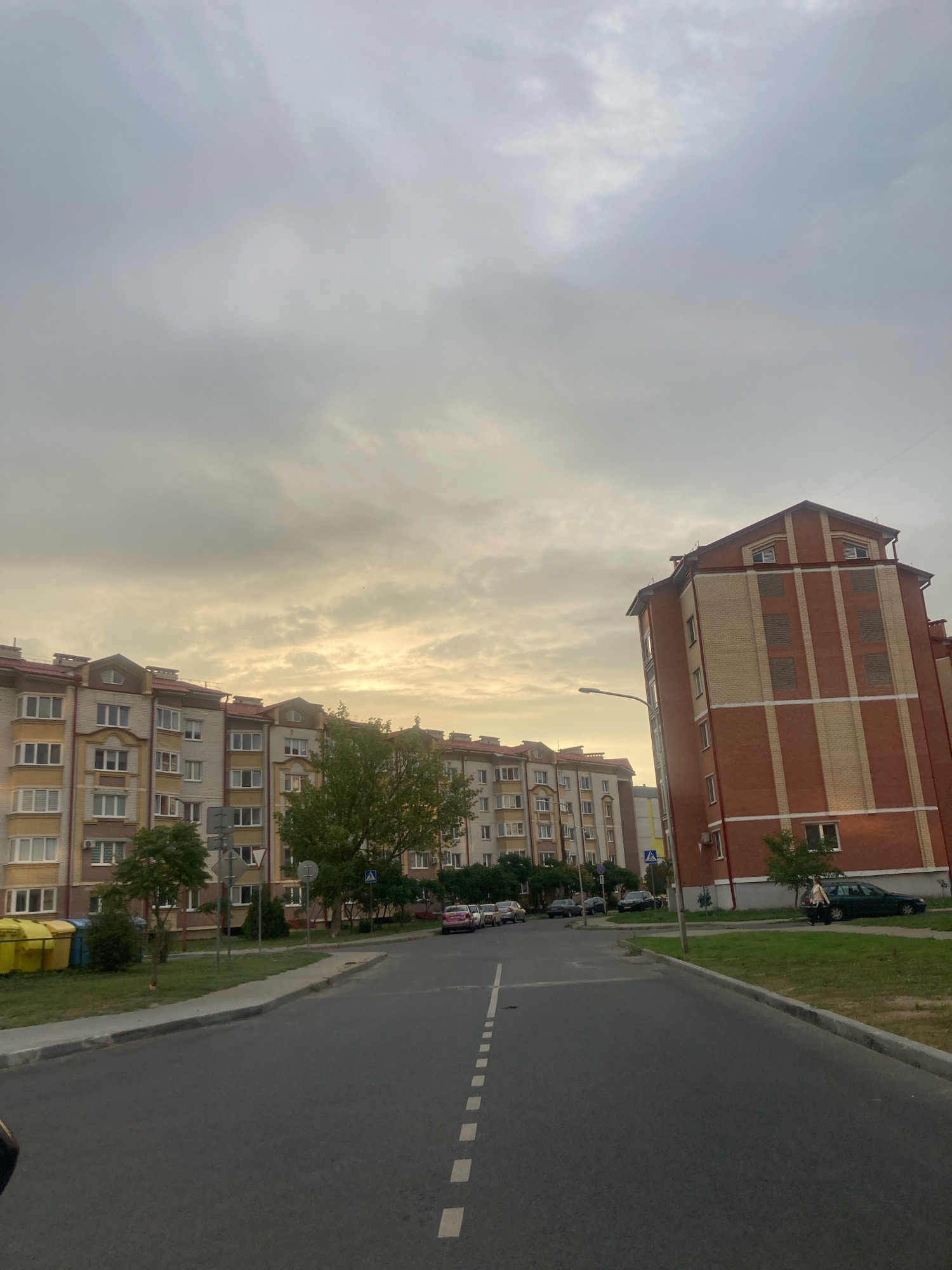 An overcast sky looms over a residential area in Brest, Belarus. The scene is dominated by two multi-story apartment buildings, one on the left with a light-colored facade and a red roof, and another on the right, a taller structure with a reddish-brown brick exterior and a similar red roof. A paved road runs through the center of the frame, with dashed white lines indicating lanes. On the left side of the road, a sidewalk borders a grassy area with several trees and two large yellow trash bins. Several cars are parked along the buildings or on the street. A person is walking on the right side of the road near the brick building. The overall impression is a typical urban residential street scene under a cloudy sky.