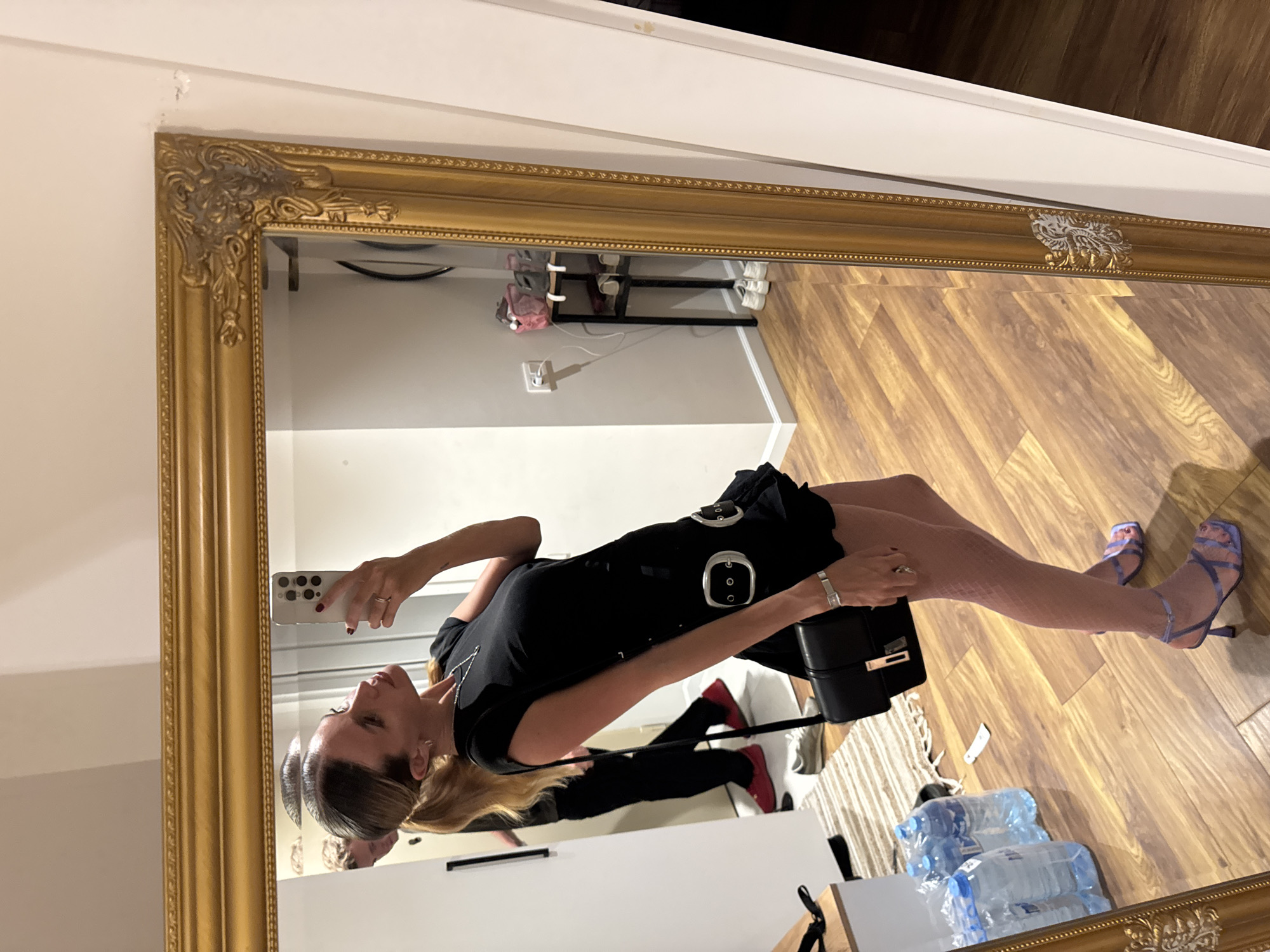 A full-length, slightly angled, vertical mirror reflects a woman taking a selfie. The woman is wearing a short, black, sleeveless dress with silver buckles and a silver bracelet on her left wrist. She is also wearing fishnet tights and light purple strappy heels. Her blonde hair is pulled back, and she is wearing subtle makeup. She holds a smartphone in her right hand to take the photo. A black handbag is visible next to her.
The reflection shows the room behind her, which appears to be an indoor setting with wood-paneled flooring and light-colored walls. A white shelf or cabinet is visible in the background, along with a black shoe rack holding a pair of white sneakers. A power outlet is also visible on the wall. Several bottles of water are placed on the floor in the lower right portion of the reflection. The lighting suggests it is daytime, but the specific time is not discernible. The overall mood seems casual and focused on the woman's attire. The mirror itself has an ornate, gold-colored frame. There is no visible text in the image.