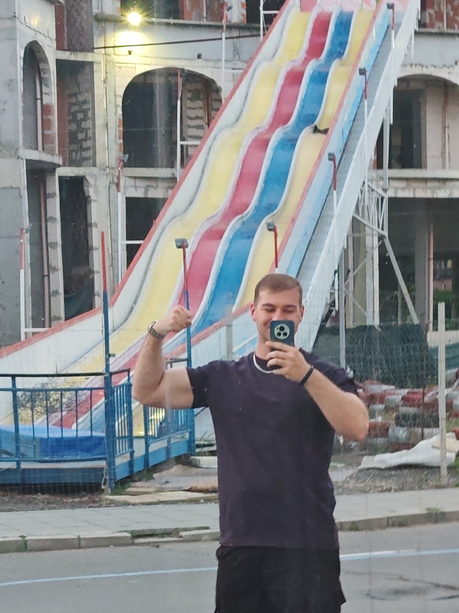 A man poses for a selfie in Nesebar, Bulgaria, with a large water slide as the backdrop. The water slide is multicolored with yellow, red, and blue sections. To the left of the slide is a partially constructed building with arched windows. The man is wearing a dark-colored T-shirt and black shorts. He flexes his arm, holding a smartphone to take a photo. In the background, there are stacked red bricks and various construction materials indicating an active building site. The scene is outside, with a paved area and street markings visible in the foreground.