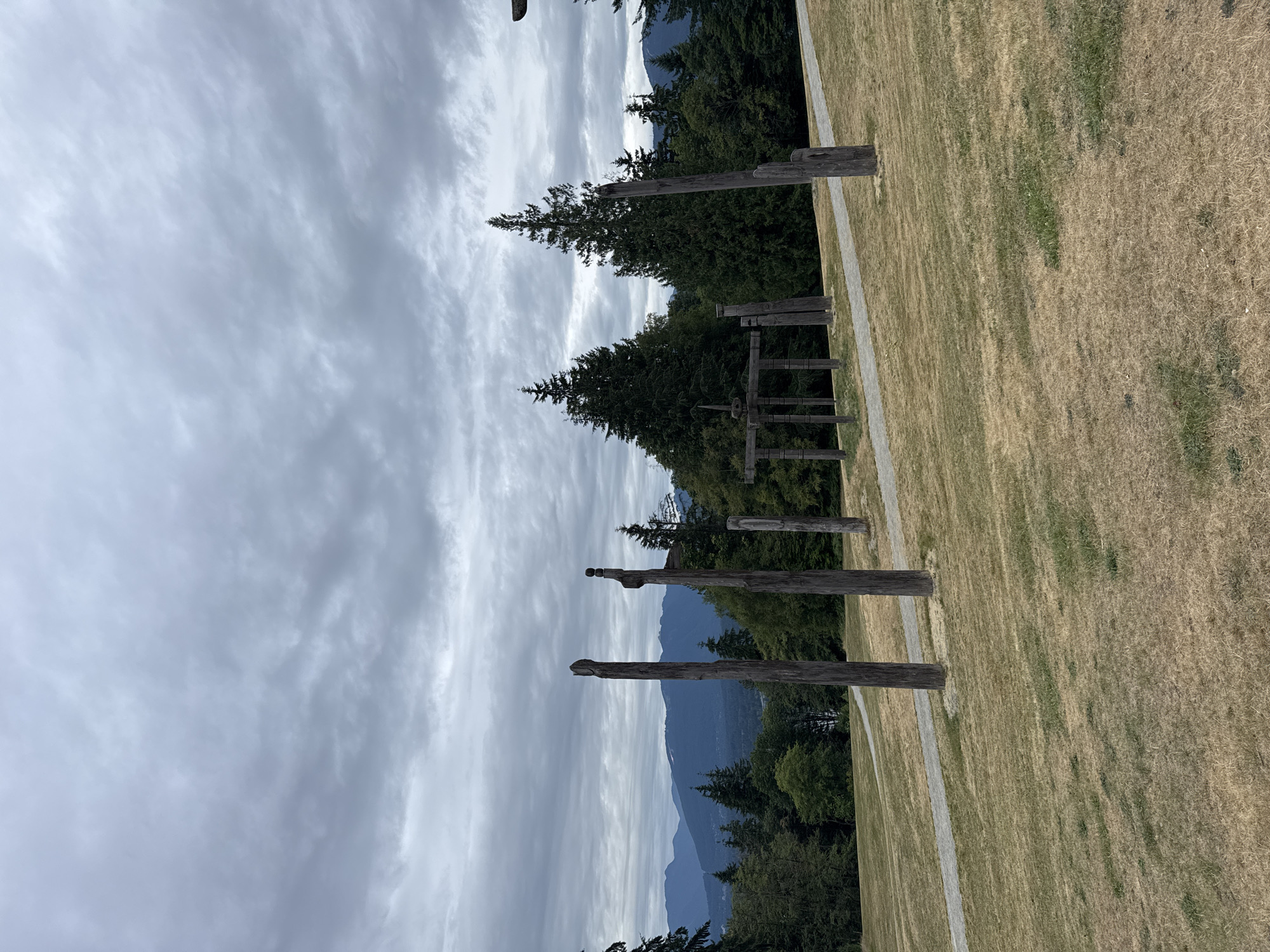 This image captures an outdoor scene at the Centennial Way Park in Burnaby, Canada, characterized by a display of totem poles and a sprawling landscape under a cloudy sky. The foreground is dominated by a dry, yellowish-brown grassy slope, indicative of late summer or early autumn. A light-colored gravel pathway curves through the grass, leading towards the center of the frame.
Scattered across the slope are several weathered wooden totem poles of varying heights and designs, some appearing to be carved, while others are simple, rough-hewn logs. One particularly prominent pole in the left foreground has a distinct, somewhat anthropomorphic top. Further into the scene, a more complex wooden structure, resembling a gate or a frame with crossbeams, stands amidst a cluster of other poles.
Behind the totem poles, a dense forest of dark green evergreen trees forms a thick backdrop. Beyond the trees, a range of blue-toned mountains stretches across the horizon, their peaks partially obscured by the overcast sky. The sky itself is filled with a layer of grey, textured clouds, creating a subdued and somewhat dramatic atmosphere. There are no people or animals visible in the image. The overall impression is one of a natural landscape with significant cultural and artistic elements.