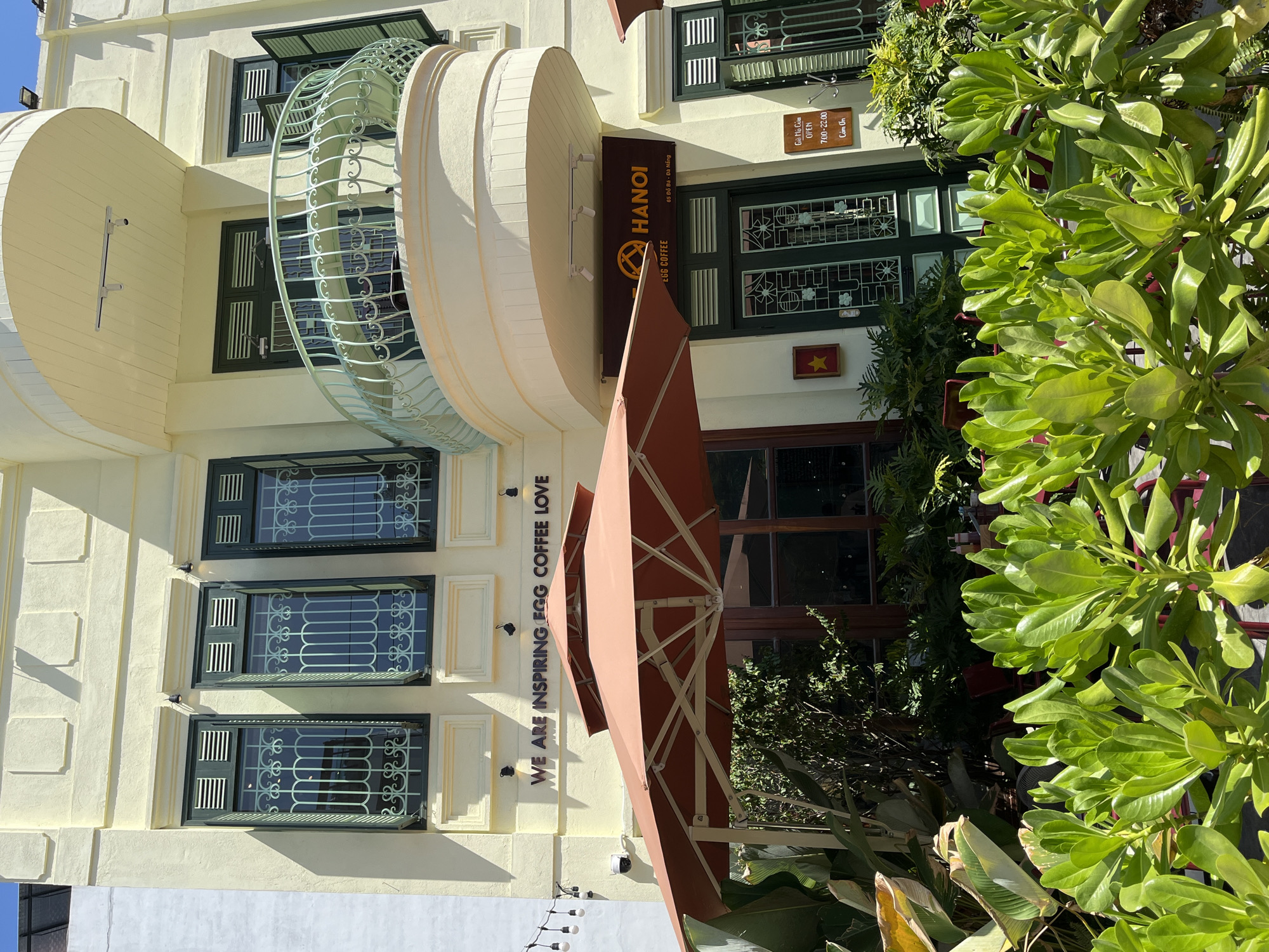 The image shows a building in Đà Nẵng, Vietnam, specifically the "Hanoi Egg Coffee" shop, located at 65 Đỗ Bá. The building is multi-storied and has a yellow façade with decorative green-framed windows. A beige-colored curved balcony is present on the upper floor. An open, peach-colored umbrella is partially visible in the foreground. Various plants surround the entrance, obscuring parts of it. A sign indicates the opening hours: 7:00-22:00. A Vietnamese flag is displayed near the entrance. Text on the building reads, "WE ARE INSPIRING EGG COFFEE LOVE." A surveillance camera is also visible.