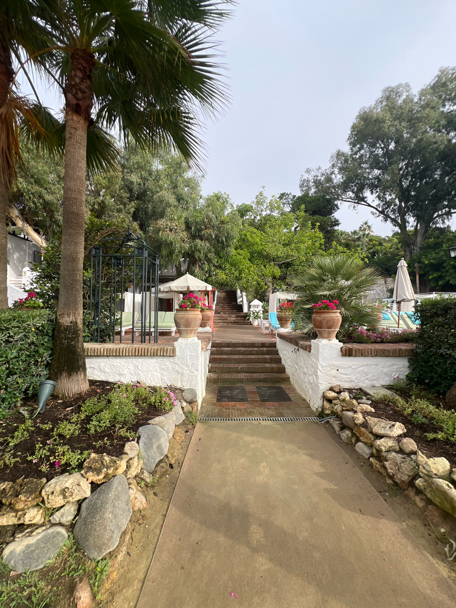 An outdoor pathway leads up a set of brick steps toward a patio area, situated amidst lush greenery and palm trees. The pathway is bordered by a stone wall and planters filled with flowers. In the background, lounge chairs and umbrellas suggest a pool or recreational area. The overall ambiance is one of a resort or upscale garden setting, likely in a location with a warm climate, such as Marbella, Spain. The sky is overcast, suggesting a potential for rain or a generally muted lighting condition. No people are visible in the image.