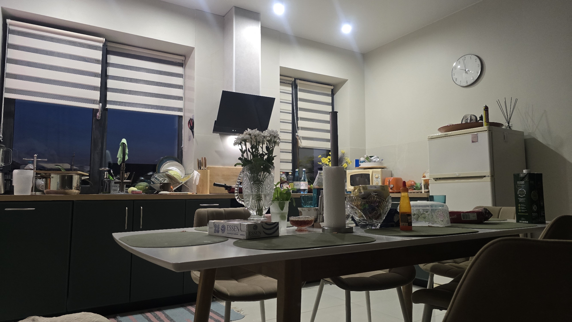 This is an indoor photograph of a kitchen and dining area, possibly in Kultaevo, Russia, given the context. The scene is lit by overhead lights and the natural light from the windows.

In the foreground, a rectangular dining table is set with several place settings, each with a dark green placemat. On the table, there are various items including a box of "ESSEN" crackers, a clear glass vase filled with white flowers, a roll of paper towels, and several bowls containing food items or decorative elements. A bottle of yellow sauce is also present. Chairs are positioned around the table, some of which are visible.

The background reveals a kitchen counter area with dark cabinetry below. On the counter, there are kitchen appliances such as a microwave, a refrigerator, and a range hood above a stovetop. Various kitchen utensils, dishes, and food items are also visible on the counter and in a drying rack. A large window with horizontal blinds is situated behind the counter, showing a dark blue twilight sky outside.

To the right of the refrigerator, a large carton of a beverage is placed. A round analog clock hangs on the wall above the refrigerator. The overall impression is of a tidy and functional living space.