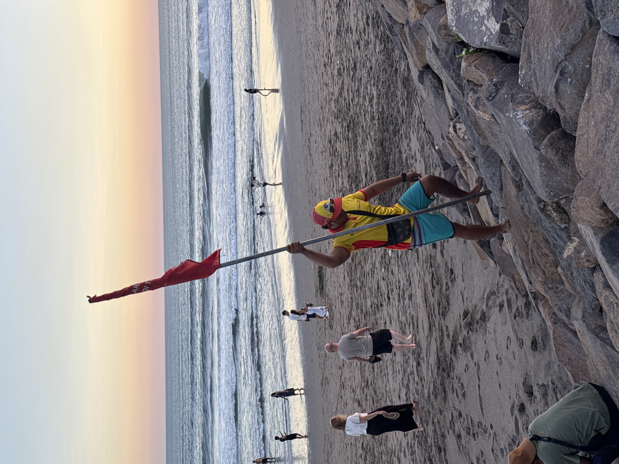 A person in a yellow shirt and blue shorts, wearing a red and yellow cap, holds a red flag on a pole on a beach in North Kuta, Indonesia. They are walking up a rocky embankment, facing away from the camera. The beach has sand and scattered rocks, with the ocean in the background. Several other people are visible on the beach and in the water. The sky is a gradient of pastel colors, suggesting either sunrise or sunset. The red flag appears to have some writing on it, but it is not legible.