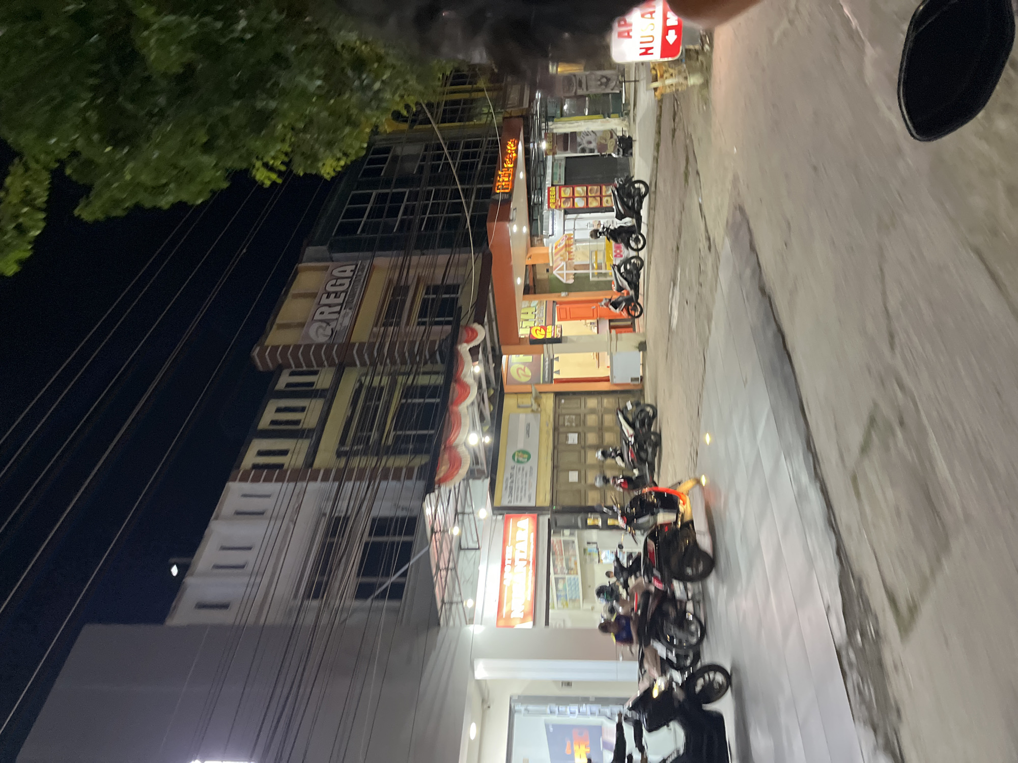The image captures a street scene at night in Langsa, Indonesia. On the left, a building with white and cream-colored facades is visible, featuring rows of windows. To its right, a yellow and brown building with vertical brick accents has a prominent sign that reads "@REGA". Further down the street, an orange-fronted building displays a sign with red lettering that appears to say "DIFI CAFE". In front of these establishments, several motorcycles are parked. A red and white sign with an arrow pointing left and the text "AP NUSAN" is visible in the lower right corner. The lighting suggests artificial illumination from streetlights and building signs. The overall environment appears to be a commercial area with buildings lining the street.