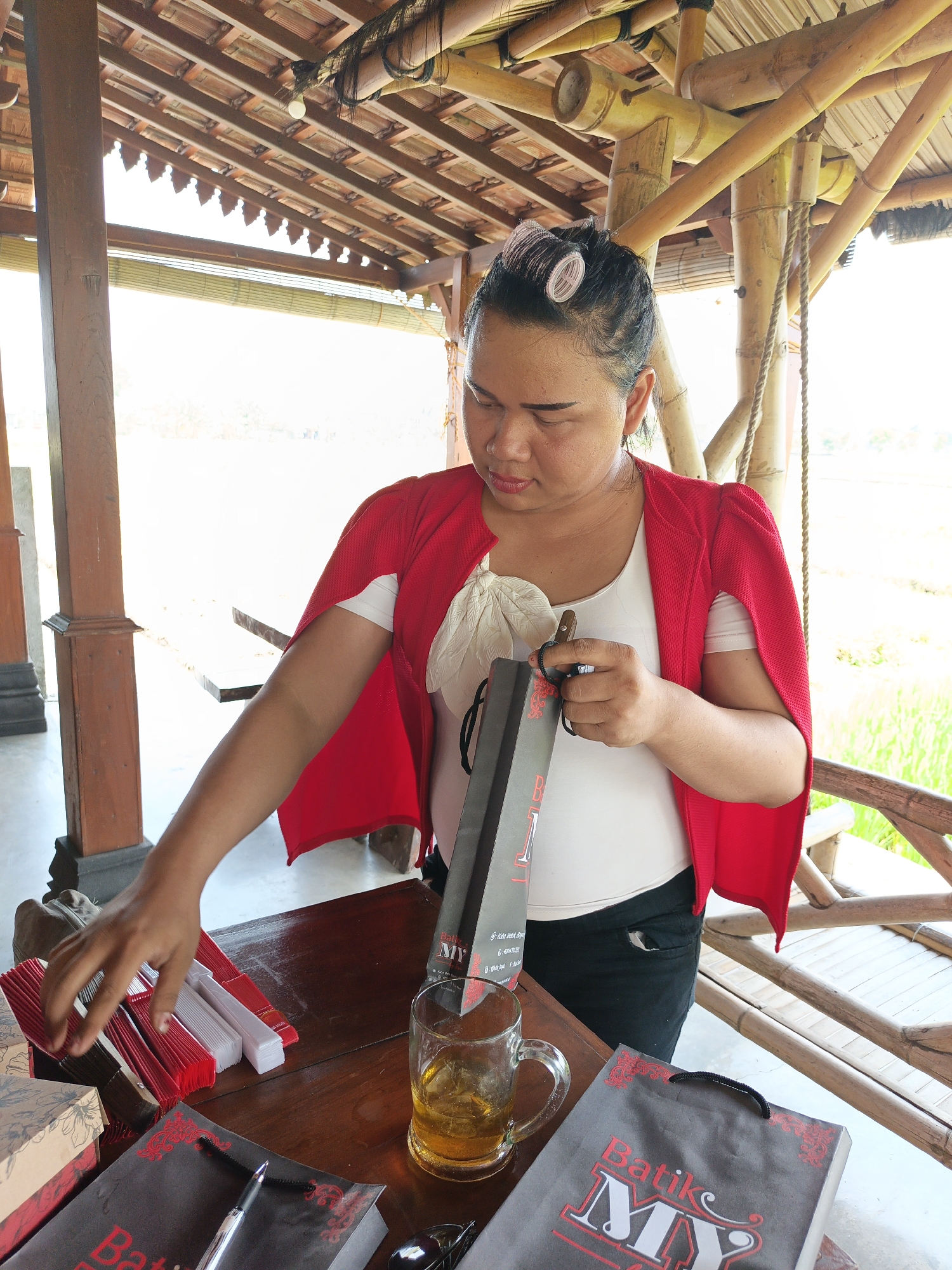 A woman with dark hair pulled back and pink curlers in it, wearing a white top and a red open cardigan, stands at a wooden table. She is holding a dark gray shopping bag with "Batik MY" printed on it in red and white lettering. In front of her on the table are several stacks of red and white folded fans, some gift boxes, a glass mug filled with a yellow beverage, and another "Batik MY" shopping bag. Her left hand is reaching towards the fans. The scene appears to be outdoors, possibly a market stall or shop, with a thatched roof supported by bamboo structures overhead. A green field is visible in the background, suggesting a rural or semi-rural setting. The image is taken from a slightly elevated angle.