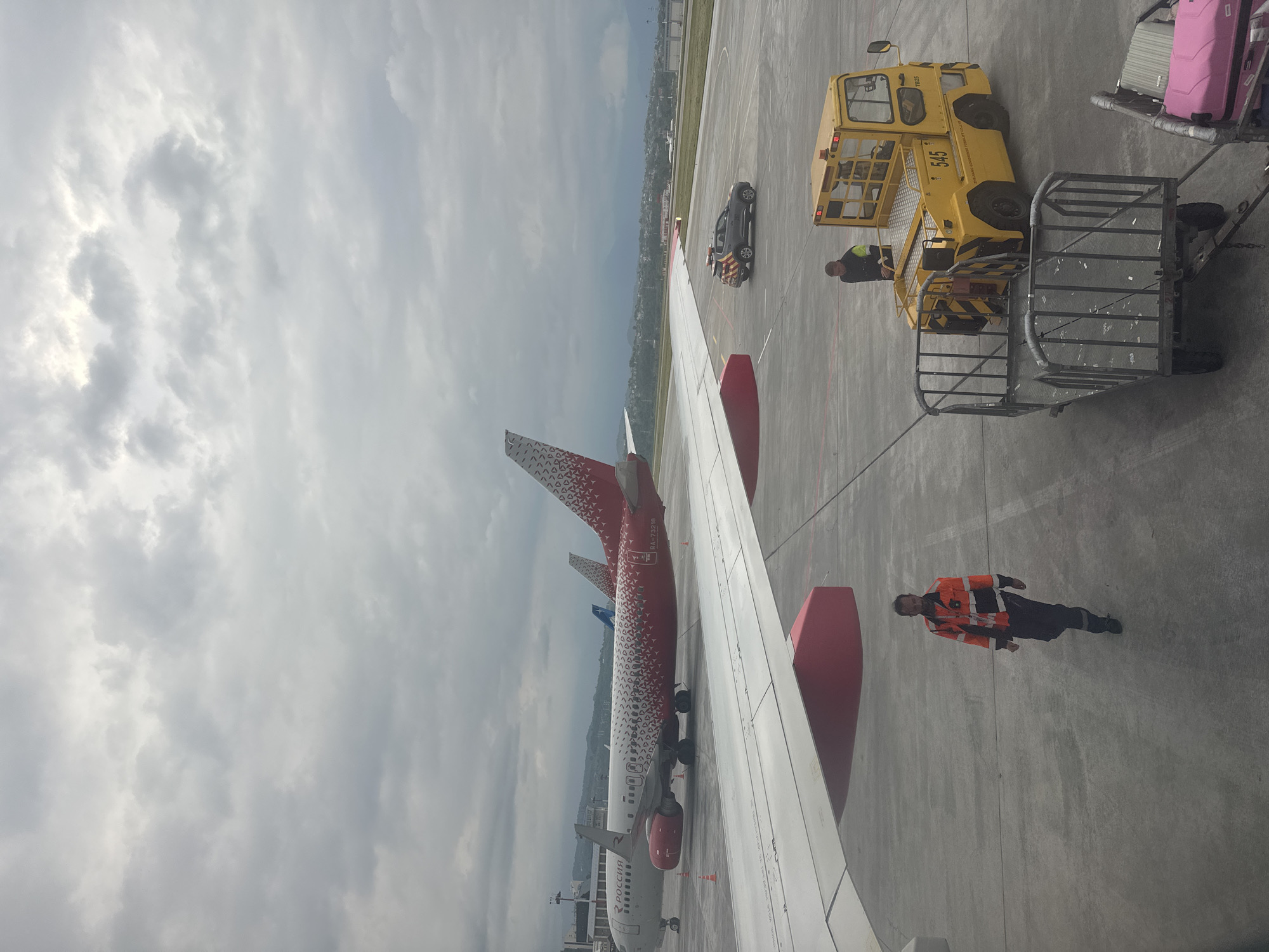 The image, taken from inside an airplane at Sochi International Airport (AER), Sochi, Russia, shows a section of the airplane wing and the tarmac below. A red and white Rossiya Airlines aircraft is visible on the tarmac. A ground crew member in a bright orange safety vest is walking on the concrete surface. A yellow aircraft servicing vehicle, likely a lift or tow tractor, is parked next to an open passenger boarding bridge. The sky is overcast with heavy clouds. Luggage is seen on the boarding bridge. A dark grey vehicle is parked near the plane's wing. The scene depicts standard ground operations at an airport.
