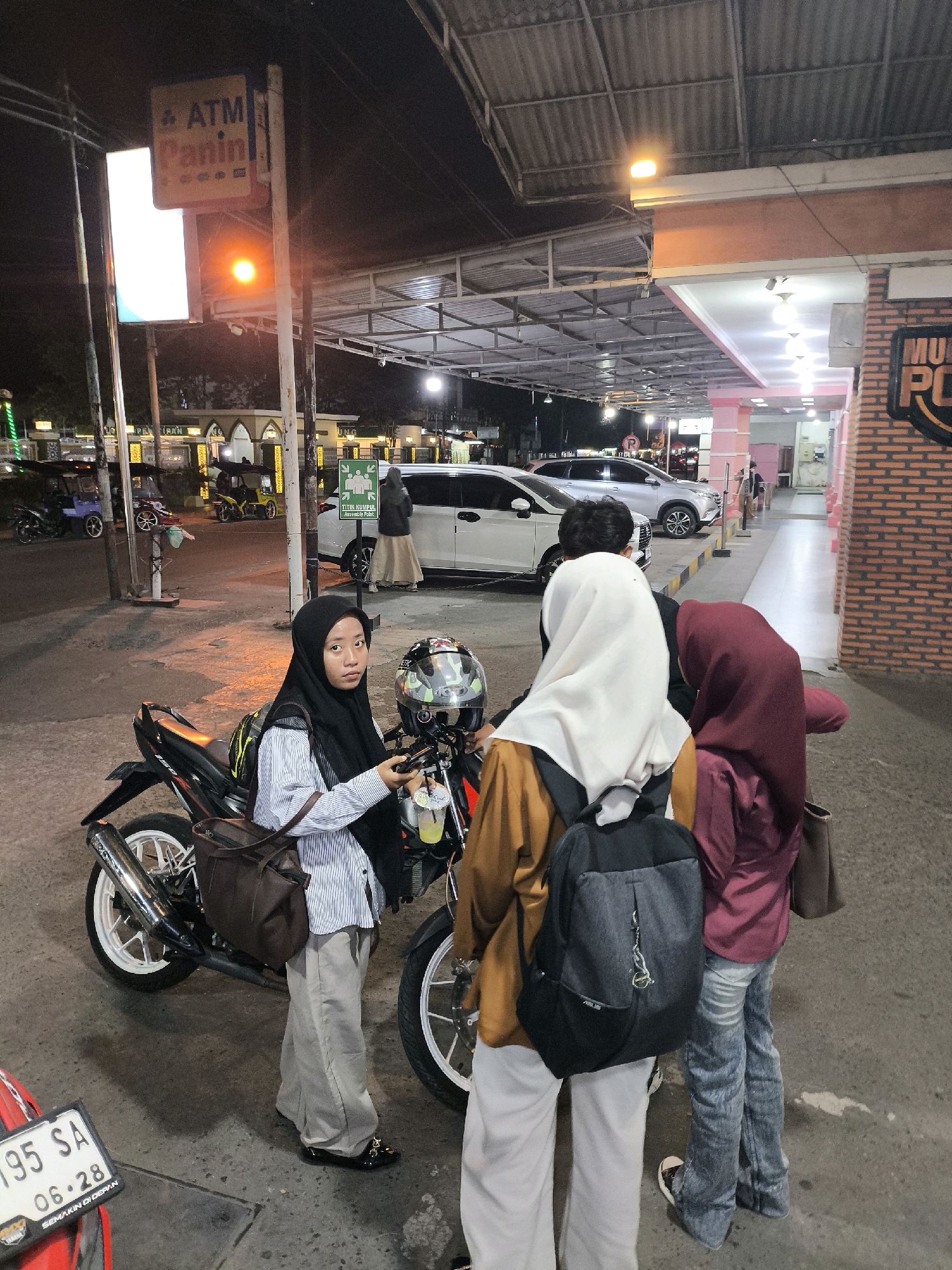 At night, in front of the Panin Bank ATM located on Jl. Nani Wartabone in Gorontalo City, Indonesia, four individuals, three women and one man, are gathered around two motorcycles. The woman in the foreground, wearing a black hijab and a striped shirt, holds a smartphone and a drink. A black helmet rests on her motorcycle. Two other women, one in a white hijab and another in a maroon hijab, stand next to her, their backs to the camera. The man, partially visible, is behind them. The street is paved and dimly lit, with the glow of streetlights and building lights illuminating the scene. Cars are parked in the background, and signage for Panin Bank ATM is visible. A red motorcycle with a license plate "195 SA 06-28" is partially in the lower left corner of the frame. The overall atmosphere is casual and social, with the group appearing to be on a break or socializing.