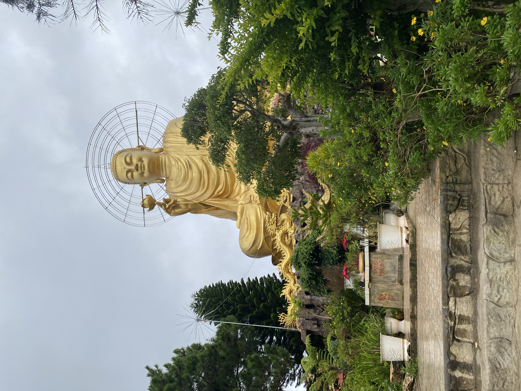 A large, golden statue of Buddha sits in a cross-legged position, holding a lotus flower in its right hand. The statue is adorned with a circular halo, and is set against a cloudy sky. The base of the statue is decorated with golden dragons and a rocky, landscaped area with trees and plants. In the foreground, there are stone steps leading up to a tiled area with potted plants and a stone monument with Vietnamese text. The scene is an outdoor religious or cultural site in Lâm Viên - Đà Lạt, Vietnam.