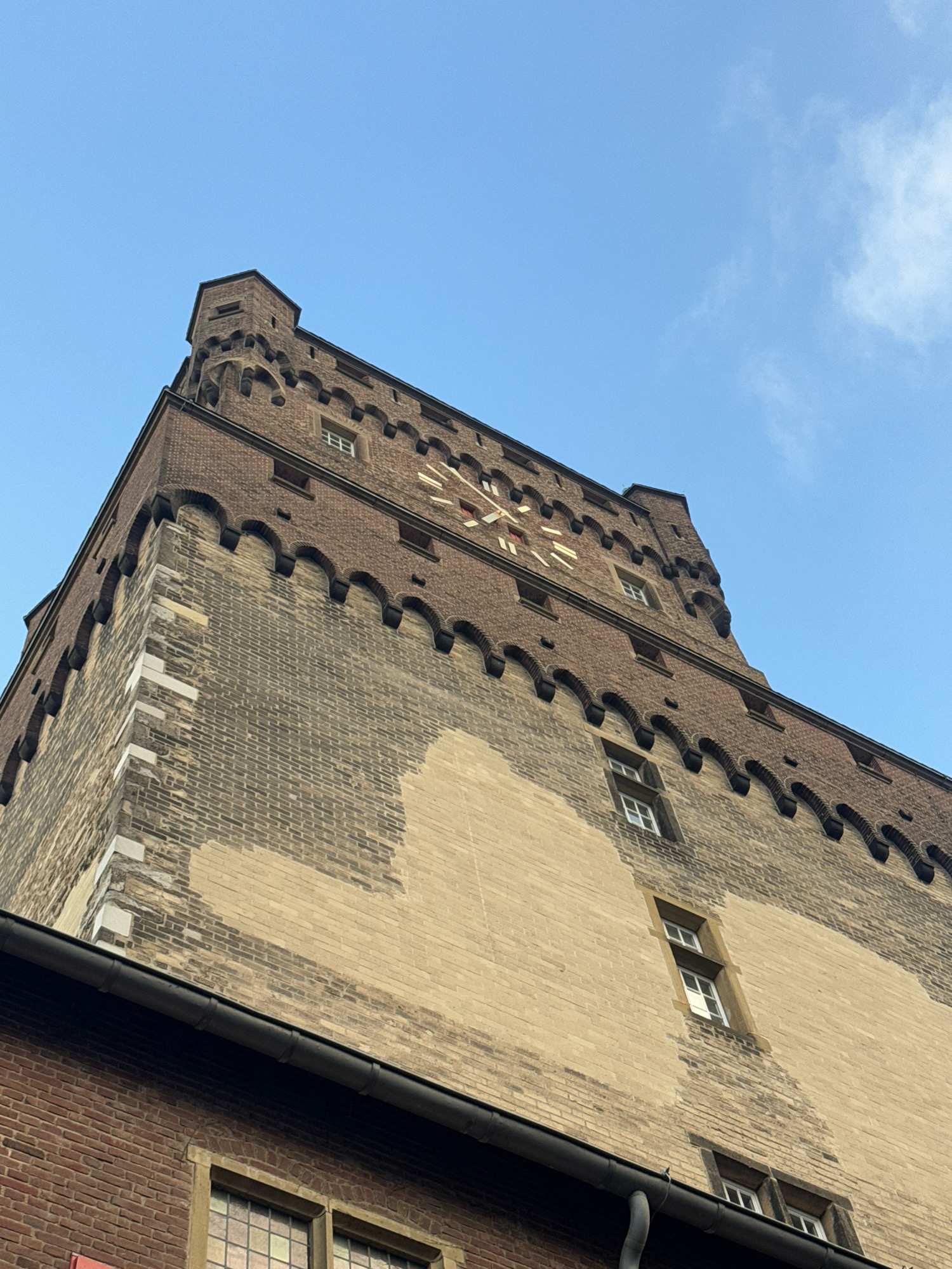 The image shows a low-angle view of the historic tower known as the "Pudding" in Cleves, Germany. The tower is constructed from brick, with sections of lighter-colored brick contrasting with darker, weathered brick. Decorative crenellations run along the upper portion of the tower, with several small windows visible on its facades. A portion of a clock face, with Roman numerals, is visible on the upper part of the tower. In the foreground, a dark gutter runs along the edge of a lower brick building. The sky above is a clear blue with a few wispy clouds. There are no people visible in the image.