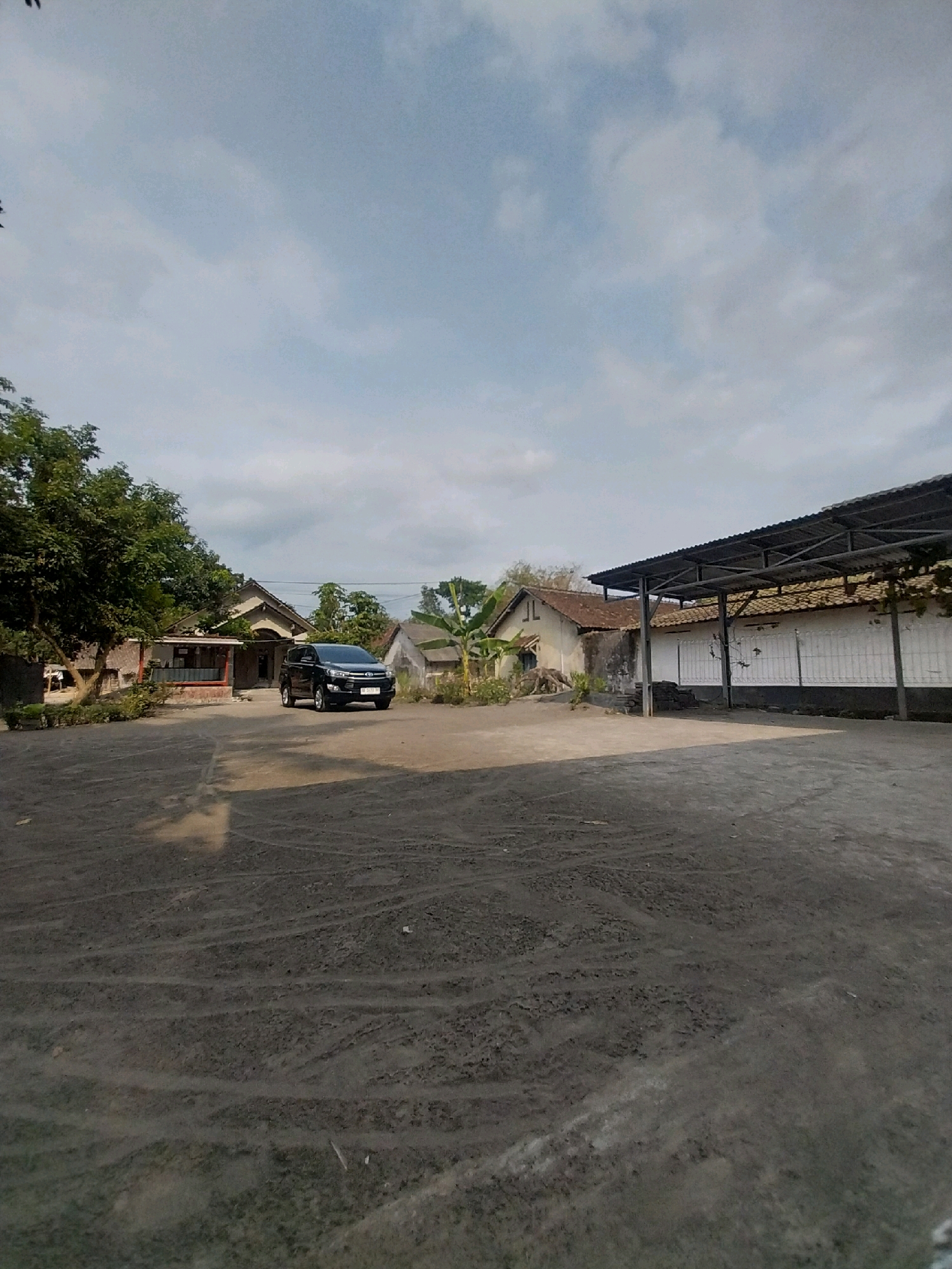 A black SUV is parked in front of a building in the city of Tijayan, Indonesia. The building appears to be a house with a red door and a tiled roof. There is a tree to the left of the building and a fence to the right. The sky is cloudy. The ground is covered in gravel.