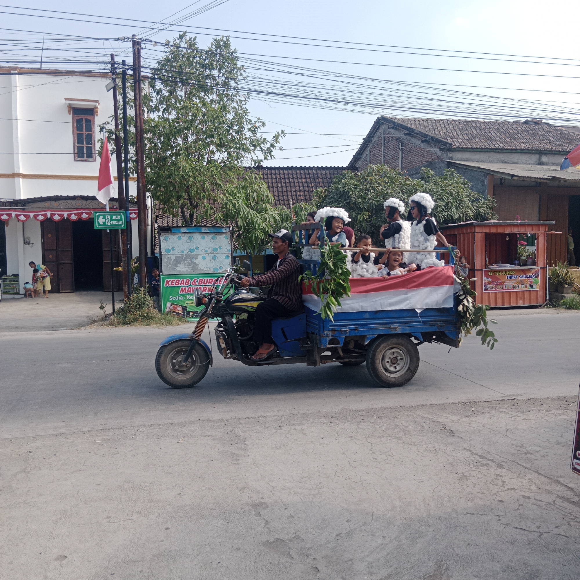In the city of Tegalrejo, Indonesia, a three-wheeled vehicle is depicted in motion on a paved road. The vehicle, a modified motorcycle with a cargo bed, is carrying several individuals, primarily young people adorned in costumes with large white, fluffy headwear and dark clothing. The cargo bed of the vehicle is draped with a flag, prominently featuring the red and white colors of the Indonesian flag.
A man, presumably the driver, is operating the vehicle, wearing a dark patterned long-sleeved shirt, dark pants, and a baseball cap. He appears to be looking forward, focused on navigating the road.
In the background, buildings with tiled roofs and white facades are visible, suggesting a residential or commercial area. A small green sign with white text and an arrow indicating "JALUR EVAKUASI" (Evacuation Route) is affixed to a pole near a building entrance, where several children and adults are gathered. Further down the street, a small kiosk or food stall with a red roof is present, displaying signage that includes text such as "ES TEH JUMBO" and "EMPAT SAUDARA". Trees with lush green foliage are scattered throughout the scene, adding to the tropical environment. Overhead, numerous power lines crisscross the sky, a common sight in many urban and semi-urban landscapes. The sky is clear and bright, indicating a sunny day. The overall impression is of a local community event or parade taking place on a sunny day in Tegalrejo.