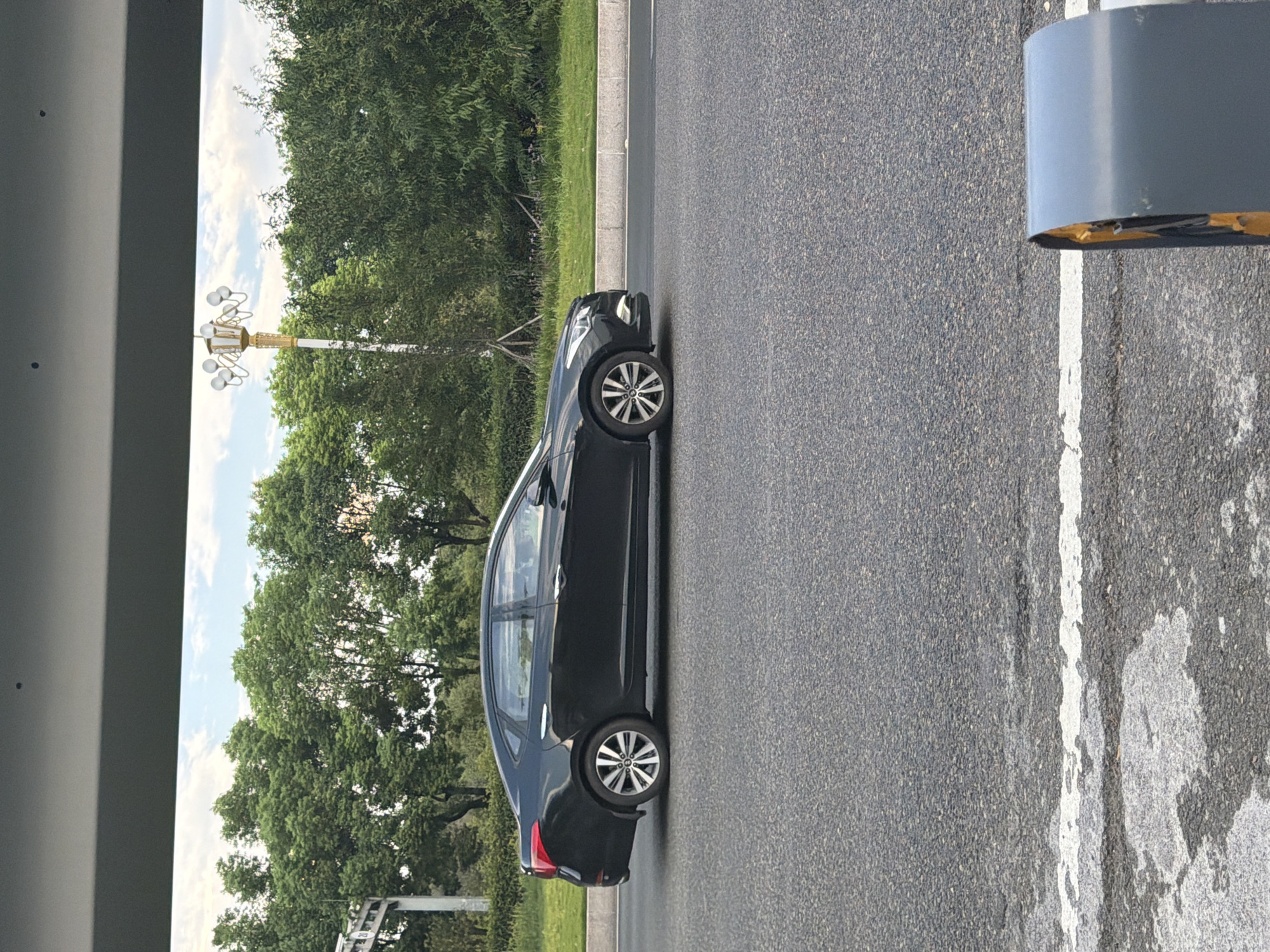 A black sedan is driving on a paved road, passing a green lawn with trees and a streetlamp. The scene appears to be in an urban or suburban area, possibly in Jiangjin, China, given the presence of lush vegetation and a style of streetlamp often seen in Asian cities. The car is captured in motion from the side view, with its headlights on. The road surface is asphalt with white lane markings. The sky is partly cloudy. In the foreground, a grey and yellow object is partially visible on the right side of the frame, possibly a traffic barrier or a piece of street furniture.