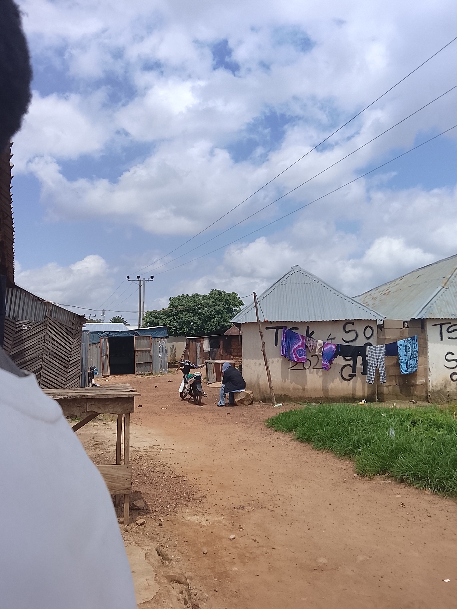 A dirt path winds through a collection of corrugated metal and wooden structures in Durumi, Nigeria. A person's white shirt is partially visible in the foreground, suggesting the perspective of someone looking out at the scene. In the mid-ground, a motorcycle is parked next to a building with a white, stucco-like wall. Laundry, including patterned pants and a blue patterned cloth, is hanging on a line suspended across the front of this building. Two individuals are present: one is seated on a rock near the laundry, and another is partially visible on the motorcycle. The building behind the laundry has a tin roof and is marked with black graffiti that appears to spell out "TSK 4 SO" and "SO G". Further to the left, another structure with a similar tin roof and open doorway stands beside a wooden table. Overhead, power lines and utility poles are visible against a partly cloudy sky, suggesting it is daytime. The overall scene depicts a typical day in a community with modest housing and infrastructure.
