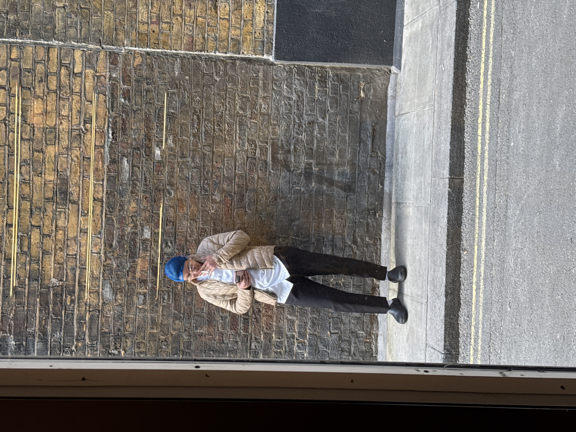 A man stands in a back alley in London, United Kingdom. He is wearing a blue beanie, a beige puffy jacket, black trousers, and black shoes. He is holding a cigarette in his right hand. He is standing in the narrow alleyway between a brick wall and a road with double yellow lines. The photograph is taken from an elevated position, looking down through a window.