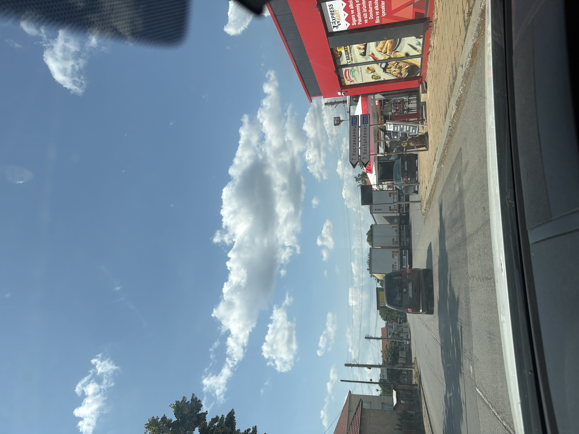 The image captures a street scene in Tsar Kaloyan, Bulgaria. The sky is blue with scattered white clouds. In the foreground, a car's dashboard is visible, suggesting the photo was taken from inside a vehicle. A brown minivan with Bulgarian license plates is in the center lane of a two-lane road. Ahead, several large white trucks are parked or moving slowly. To the right, a bright red building with a sign for "EXPRESS MART" is prominent, featuring images of food on its windows. A smaller sign on the building reads "Sigara ve alkol satisi" in Turkish, meaning "cigarette and alcohol sales." Also visible are directional signs pointing towards "Kostandenec" (likely Kyustendil, a city in Bulgaria) with the number 2002. The street is paved, with sidewalks and some vegetation visible. A few people are sparsely present near the mart. The overall impression is a roadside stop in a Bulgarian town on a sunny day.