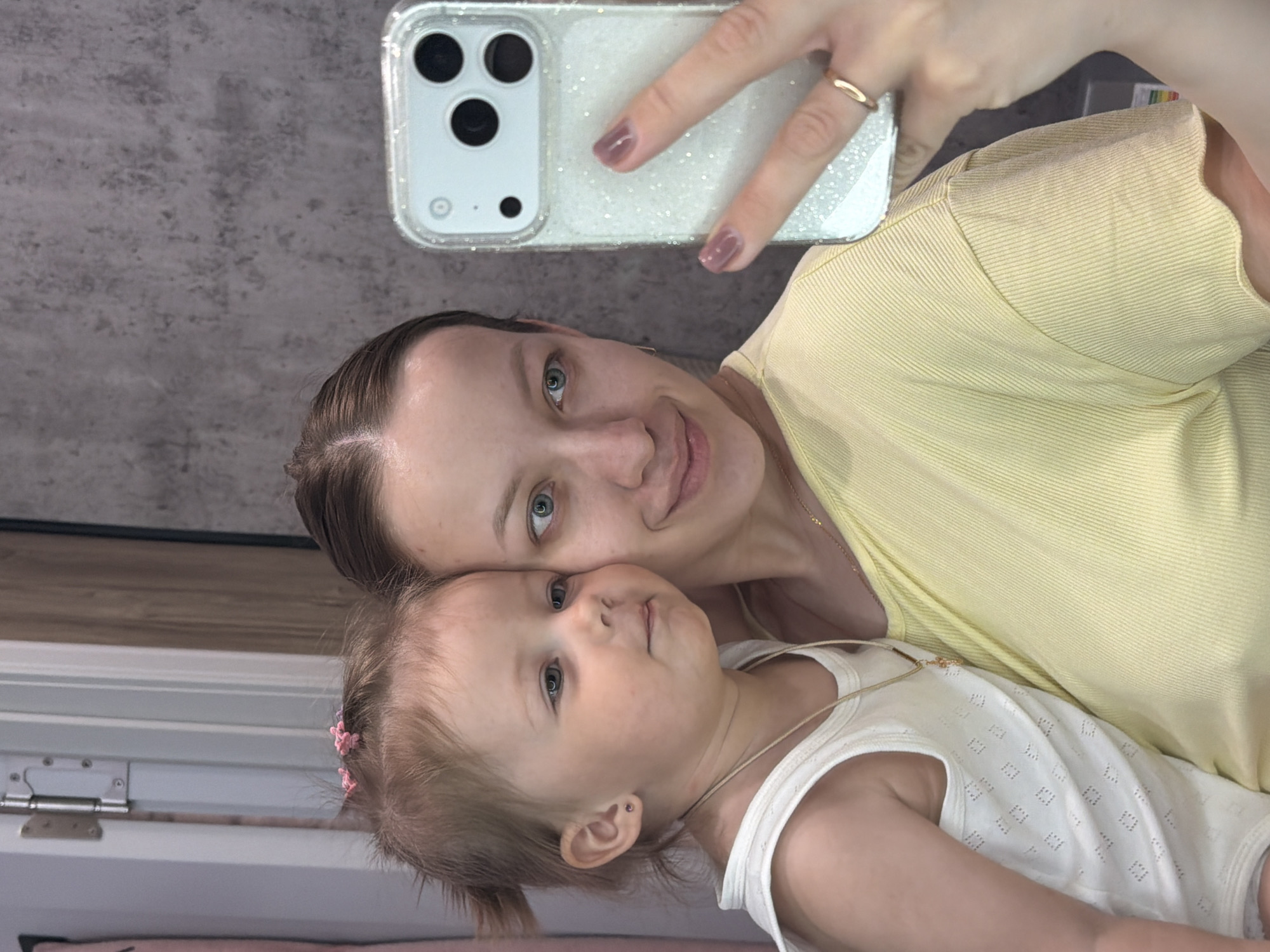 A woman is taking a selfie with a toddler. The woman has blue eyes, light brown hair pulled back, and is wearing a yellow t-shirt and a gold necklace. She is holding a white phone with a glittery case in her right hand, showing a gold ring on her finger and light brown nail polish. The toddler is wearing a white top and a gold necklace. A small pink hair clip is visible in the toddler's hair. The background includes a gray wall and a white door frame. The location is not discernible from the image.