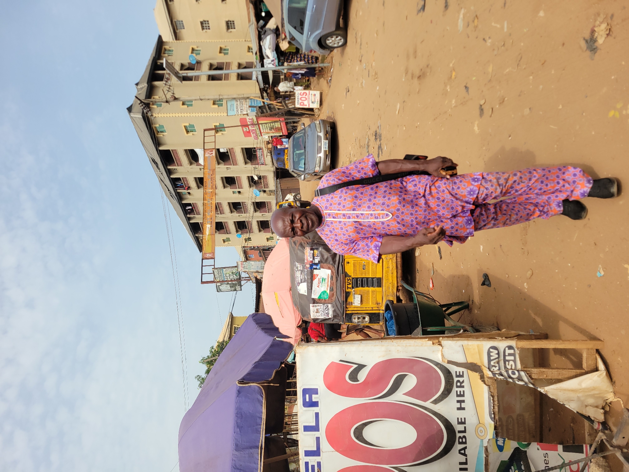 The image shows a man standing in what appears to be a bustling street scene in Nkpor, Nigeria. He is wearing a patterned purple shirt and trousers with black shoes and has a bag slung over his shoulder. Behind him is a yellow auto rickshaw (Tuk-Tuk) with various items attached to it, including stickers and a black bin. In the foreground, there is a tattered sign advertising "ELLA OS," with a faded awning above it. The ground is dirt and debris-strewn. Buildings of multiple stories are visible in the background, along with other signs and cars. The overall impression is of a busy, somewhat chaotic urban environment. Several signs are visible, advertising a church of Pentecost, ADAMAZI POS, and GOLDEN EAGLE. The OCR text is visible on the sign and also shows tvs and acycors logos.