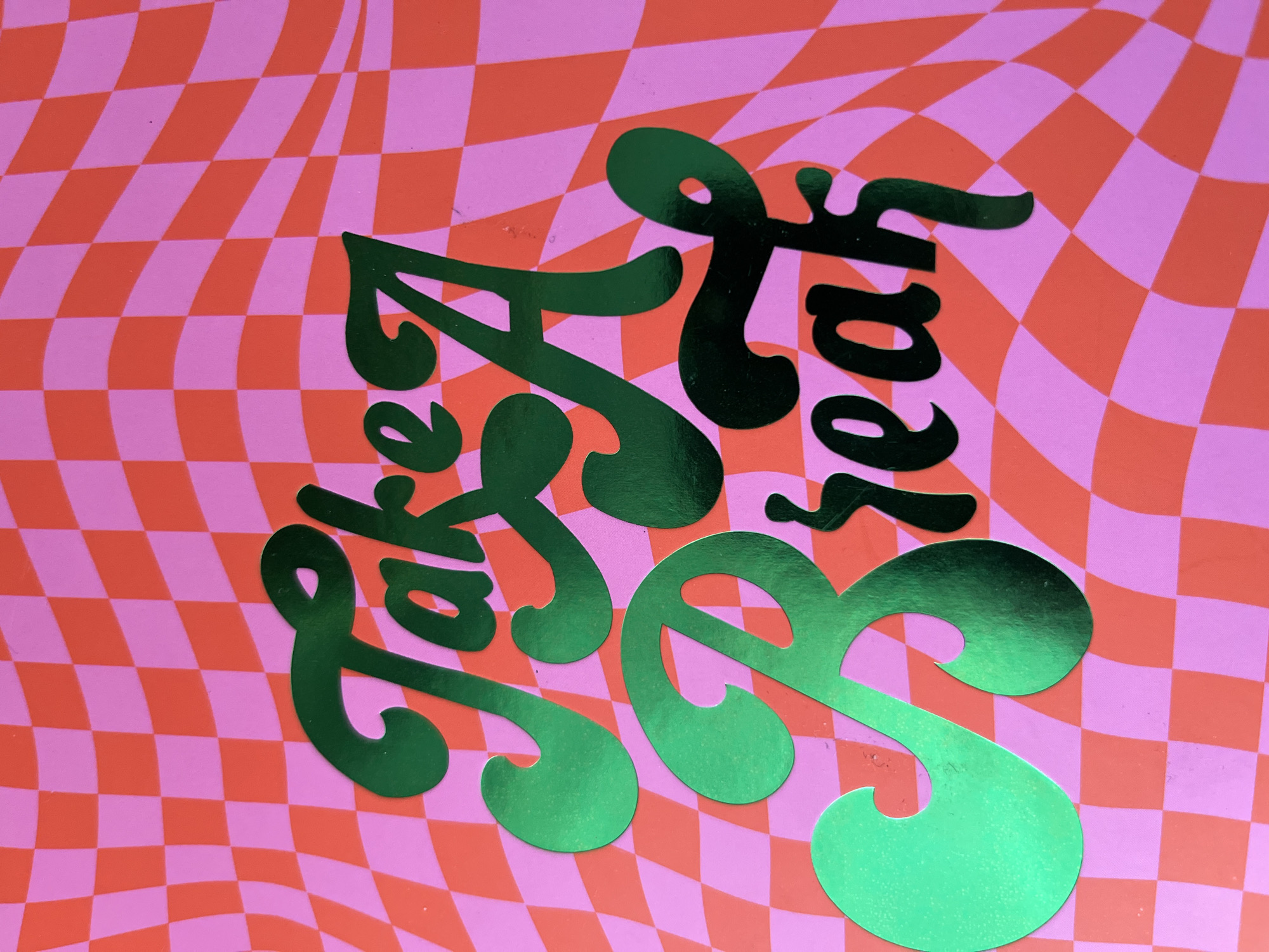 The image displays a vibrant graphic design featuring a distorted checkerboard pattern as its background. The checkerboard consists of bright pink and reddish-orange squares that ripple and undulate, creating a wavy, psychedelic effect.
Overlaid on this background is text in a metallic, shimmering green color. The text is rendered in an elaborate, flowing cursive script and is oriented diagonally across the frame, reading from the lower left towards the upper right. The phrase appears to be "Life is a Beach." The word "Life" begins with a large, stylized 'L', while the word "Beach" starts with an equally prominent and ornate capital 'B' that curves dramatically. The metallic green text reflects light, causing variations in its hue from darker green to lighter, almost lime green, suggesting an iridescent or holographic material like foil or vinyl.
There are no people or specific activities depicted. The scene is static, focusing entirely on the graphic design, which likely serves as a poster, sign, or part of a product. The overall aesthetic is playful and retro, evoking a cheerful mood. The surface shows minor textural imperfections and reflections, indicating it is a photograph of a physical object.