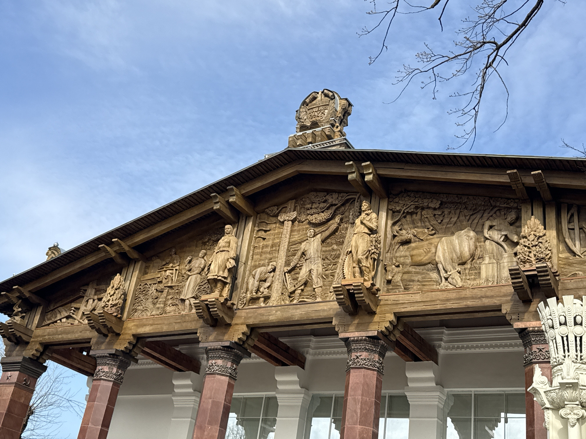 The image depicts a portion of the façade of a building, specifically the former Belarusian SSR pavilion at the VDNKh exhibition center in Moscow, Russia. The building features a high, intricately carved wooden frieze depicting scenes of agricultural and industrial labor, including farmers, loggers, and livestock. The frieze is supported by a series of columns, the lower portions of which are a reddish-brown color and the upper portions of which are white. The roof is visible above the frieze, and the building is capped with a decorative element. The sky is visible in the background, partially obscured by bare tree branches. The overall style is Soviet monumentalism, characterized by its grand scale and celebratory depiction of collective labor. The building's windows and the surrounding environment suggest it is daytime.