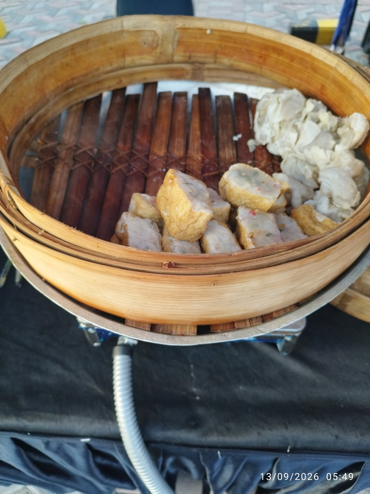 A close-up, overhead shot showcases a bamboo steamer filled with food. The steamer, constructed from light brown bamboo rings, contains what appears to be pieces of fried tofu and some white, shredded food, possibly steamed dumplings. The food is arranged in the center of the steamer, which rests on a metal rim. Below the steamer, a black cloth is visible, and a silver, corrugated hose extends upwards from the bottom of the frame, connecting to an unseen apparatus. The background is blurred, but suggests an outdoor setting with a patterned surface and a hint of a blue object in the upper right. A timestamp "13/09/2026 05:49" is present in the bottom right corner of the image. The scene is likely set in Mojokerto, Indonesia, possibly at a street food stall or a market. No people are visible in the frame.