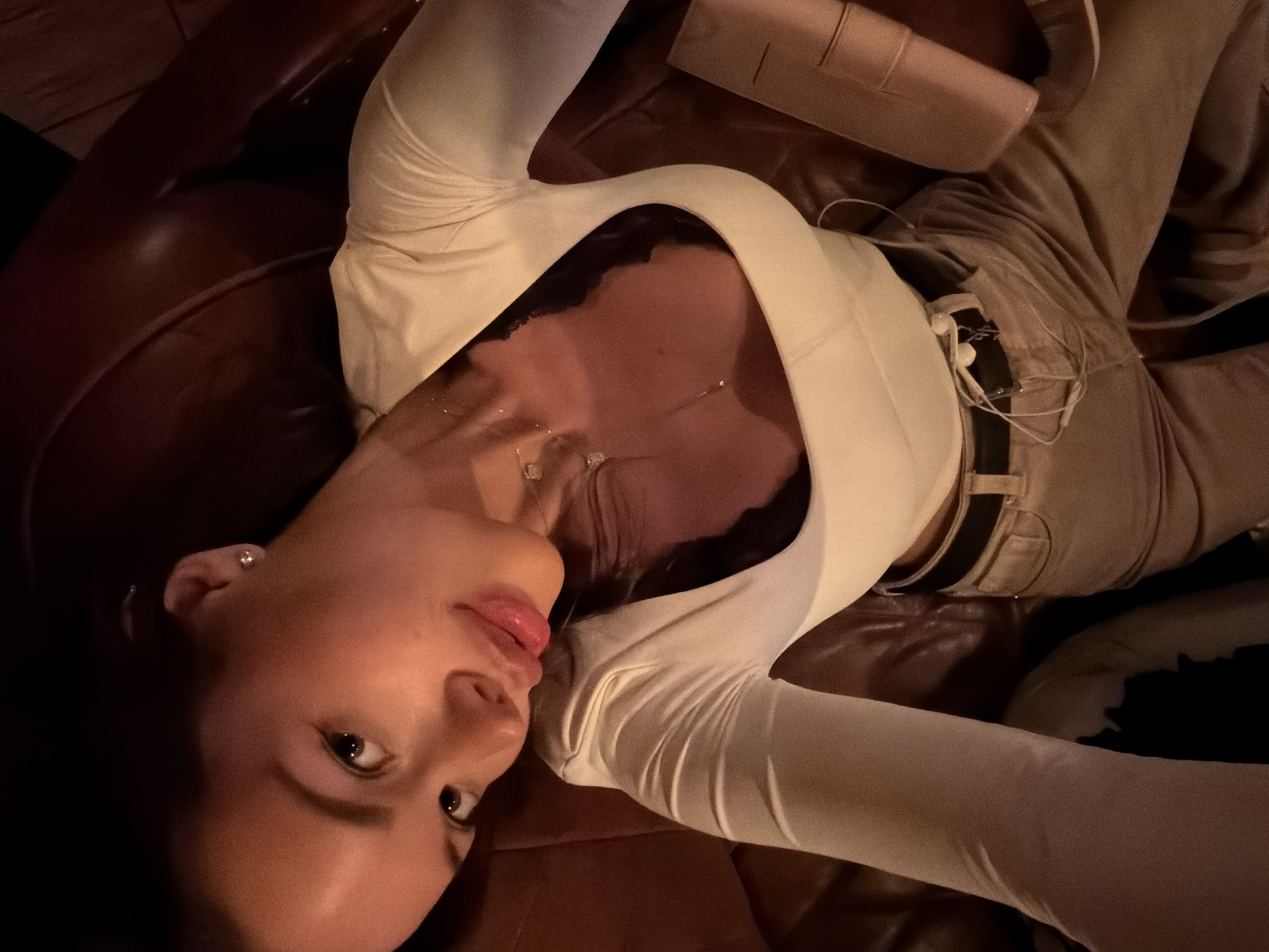 A young woman is depicted in a medium shot, lying on a dark brown, possibly leather, couch or armchair. She is positioned diagonally across the frame, with her head turned towards the viewer. She wears a long-sleeved, light-colored (cream or off-white) top with a wide neckline, revealing a necklace with a small pendant on her chest. Her attire also includes light brown or beige pants with a dark belt. White wired earphones are visible hanging near her waist. Near her head, a light pink or nude-colored rectangular clutch bag or wallet rests on the furniture.
The setting appears to be indoors, likely an evening or nighttime scene, characterized by dim, warm ambient lighting that creates reflections on the glossy surface of the furniture and her skin. Her expression is calm, with her lips slightly parted. The photograph captures her in a posed moment, looking directly at the camera. No visible text is present in the image. The location context provided is Moscow, Russia.