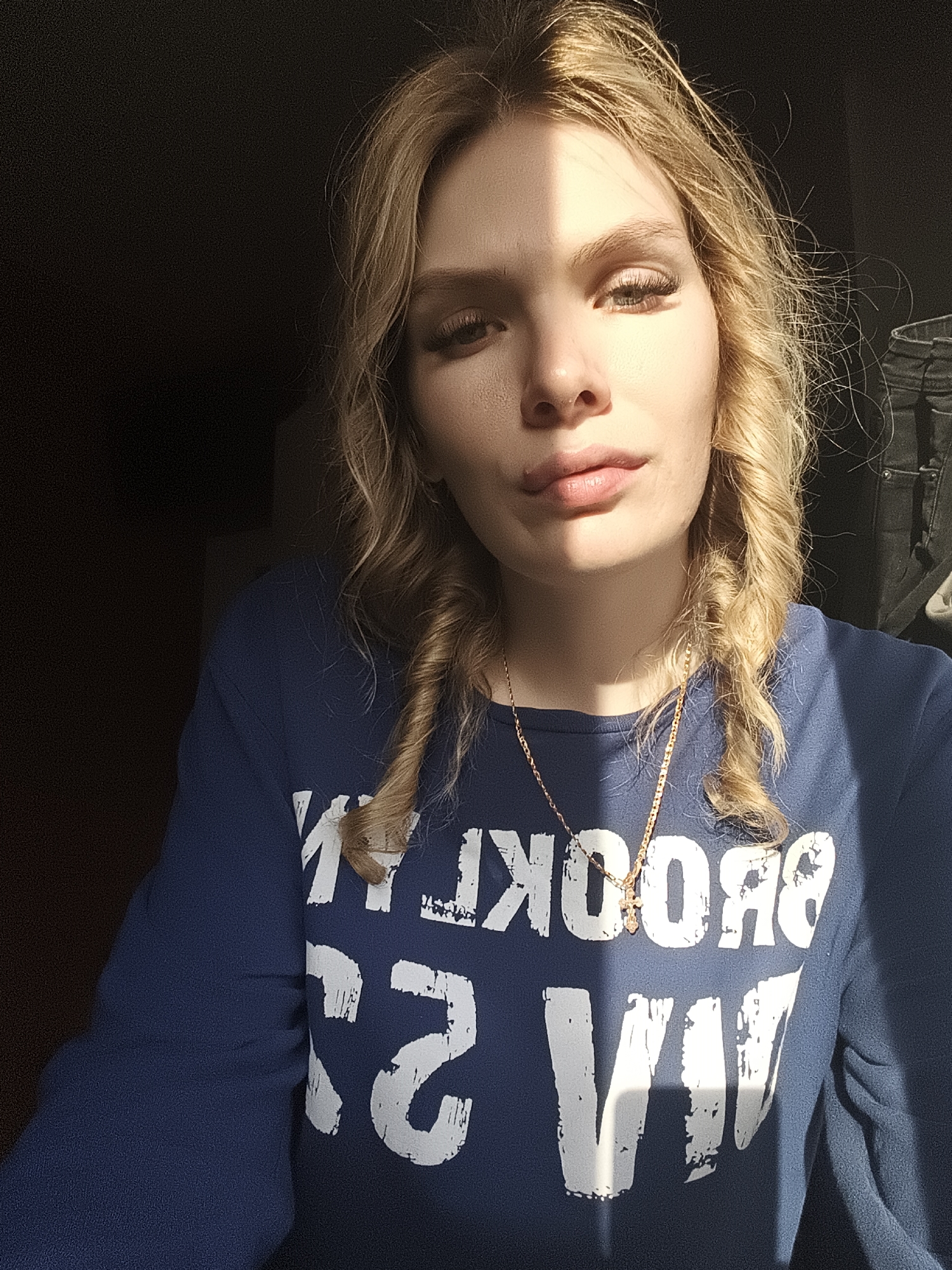 A fair-skinned woman is centered in a selfie shot, wearing a dark blue sweatshirt with the word "BROOKLYN" printed in white letters. She has blonde, wavy hair, braided into two pigtails. A gold necklace with a cross pendant hangs around her neck. The lighting is dramatic, with a stark line of sunlight illuminating half of her face. The background is dark and indistinct, but a piece of dark clothing is visible behind her right shoulder. The location, Русский Вожой, Russia, is not identifiable in the image.