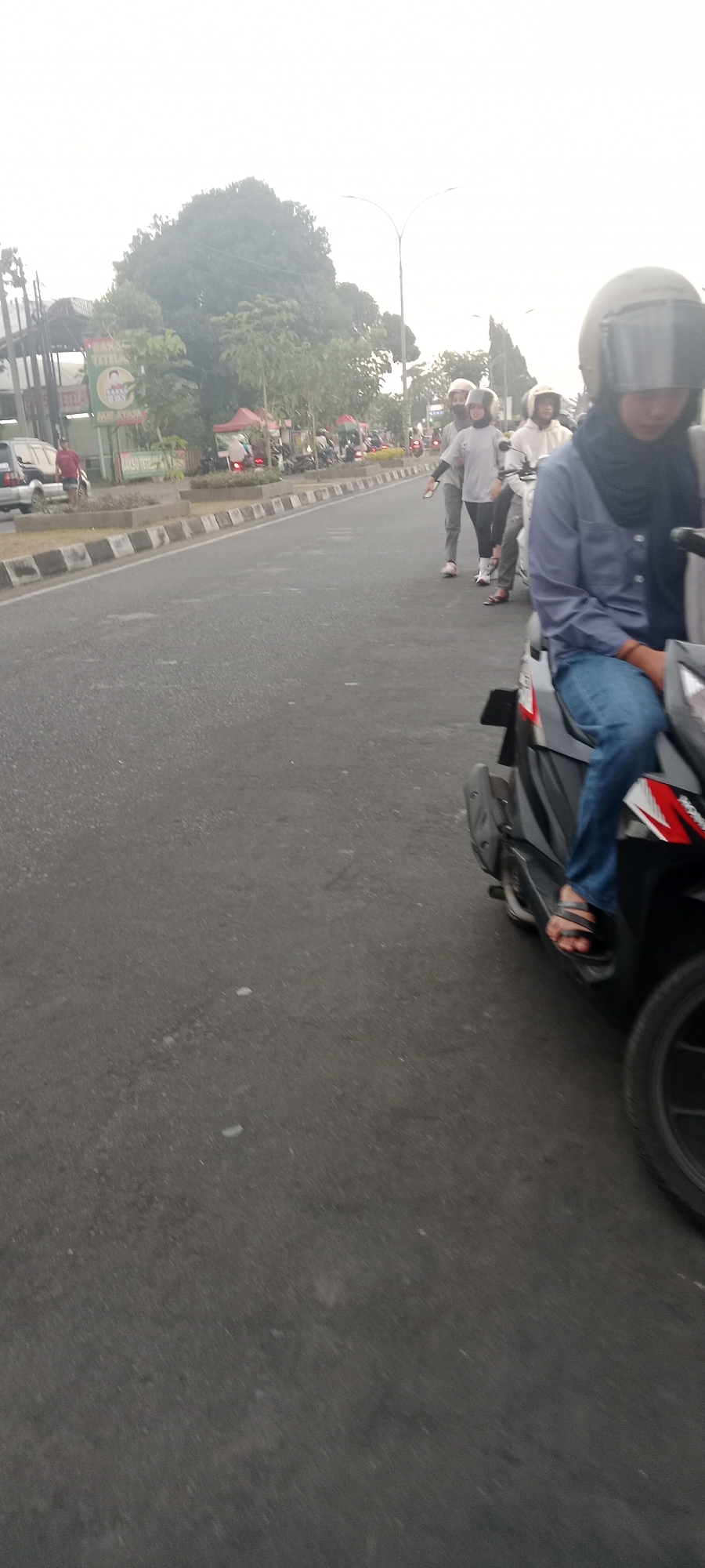 In Ngemplak, Indonesia, on a hazy day, several people are riding motorcycles along a paved road with a decorative median. In the foreground, a person wearing a blue long-sleeved shirt, blue jeans, and sandals rides a black motorcycle, their face obscured by a helmet. To the left, a dark-colored car is parked near a building with banners. Further down the road, several other individuals on motorcycles are visible, some wearing helmets and casual clothing. The environment appears to be an urban or suburban area with trees and buildings lining the street. The road surface is asphalt with white and black markings on the median.