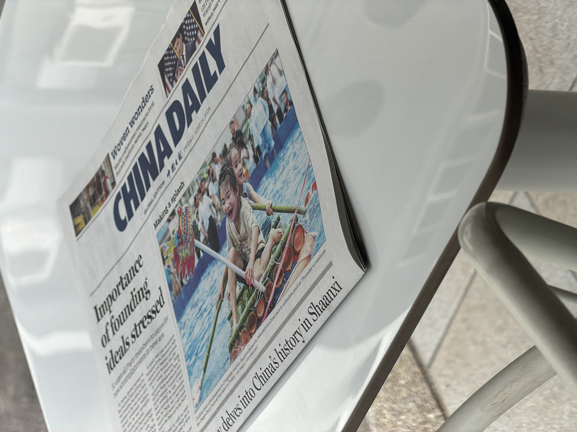 The image shows a close-up, overhead view of a newspaper on a white table. The newspaper is identified as "CHINA DAILY" and the date is Friday, June 13, 2026. The headline across the top of the newspaper reads "Importance of founding ideals stressed." Below this, a large photograph depicts children participating in a dragon boat race. Another headline on the left side of the newspaper states, "delves into China's history in Shaanxi." The newspaper is positioned at an angle on the table, with the right side of the table and a chair arm visible in the foreground. The lighting suggests an indoor setting, possibly a cafe or restaurant, with natural light reflecting on the table's surface. There is no identifiable information within the image to indicate the location is Bangkok, Thailand.