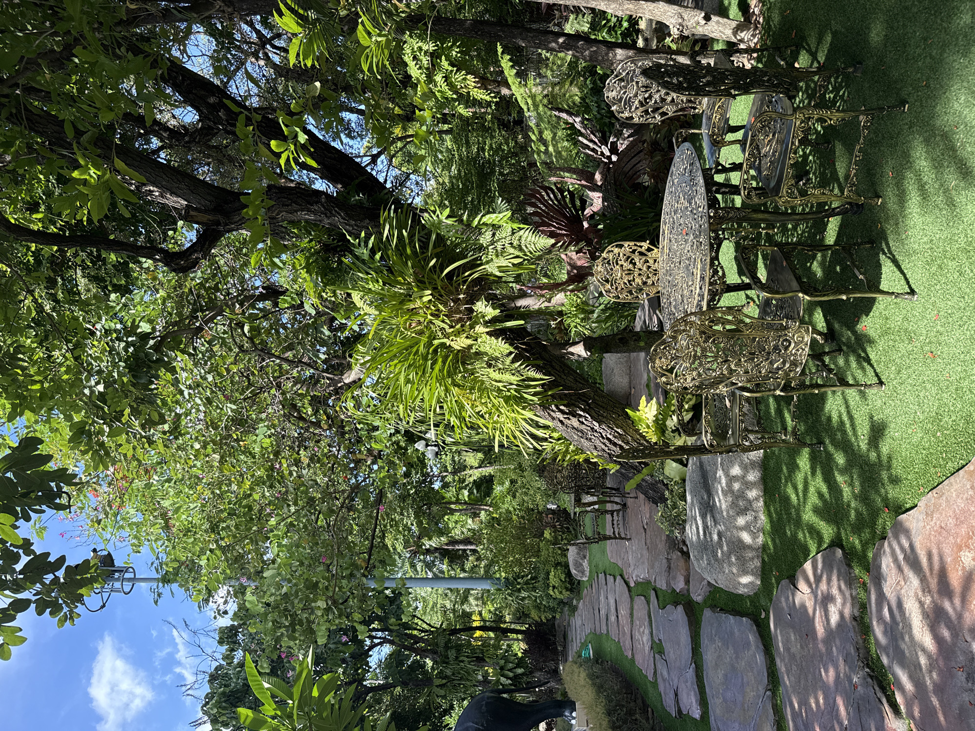 This photograph captures a tranquil outdoor scene in Samrong Nuea Subdistrict Municipality, Thailand. A set of ornate, antique-style metal outdoor furniture, including a round table and four chairs, is arranged on a patch of green turf beside a stone-paved pathway. The path meanders through a lush garden setting, densely populated with trees, ferns, and various plants, creating a shaded and verdant environment. A large tree with epiphytic plants growing on its trunk is prominently featured near the furniture. The sky is partially visible through the tree canopy, revealing a few clouds. A lamp post and the hint of a statue add to the character of the scene.