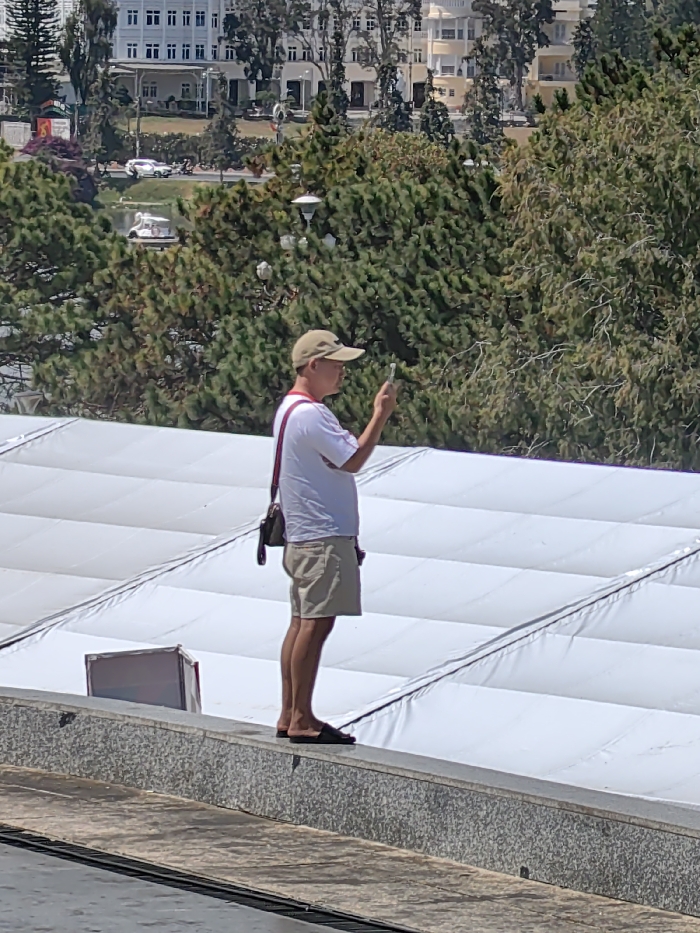A man stands on a concrete ledge, holding a phone. He is dressed in a white shirt, khaki shorts, a tan baseball cap, and sandals. A shoulder bag hangs from his left shoulder. Behind him, a large white structure stretches out, possibly a tent or inflated building. The background features trees, a body of water with a boat, buildings, and a clear sky. The scene is bathed in sunlight.