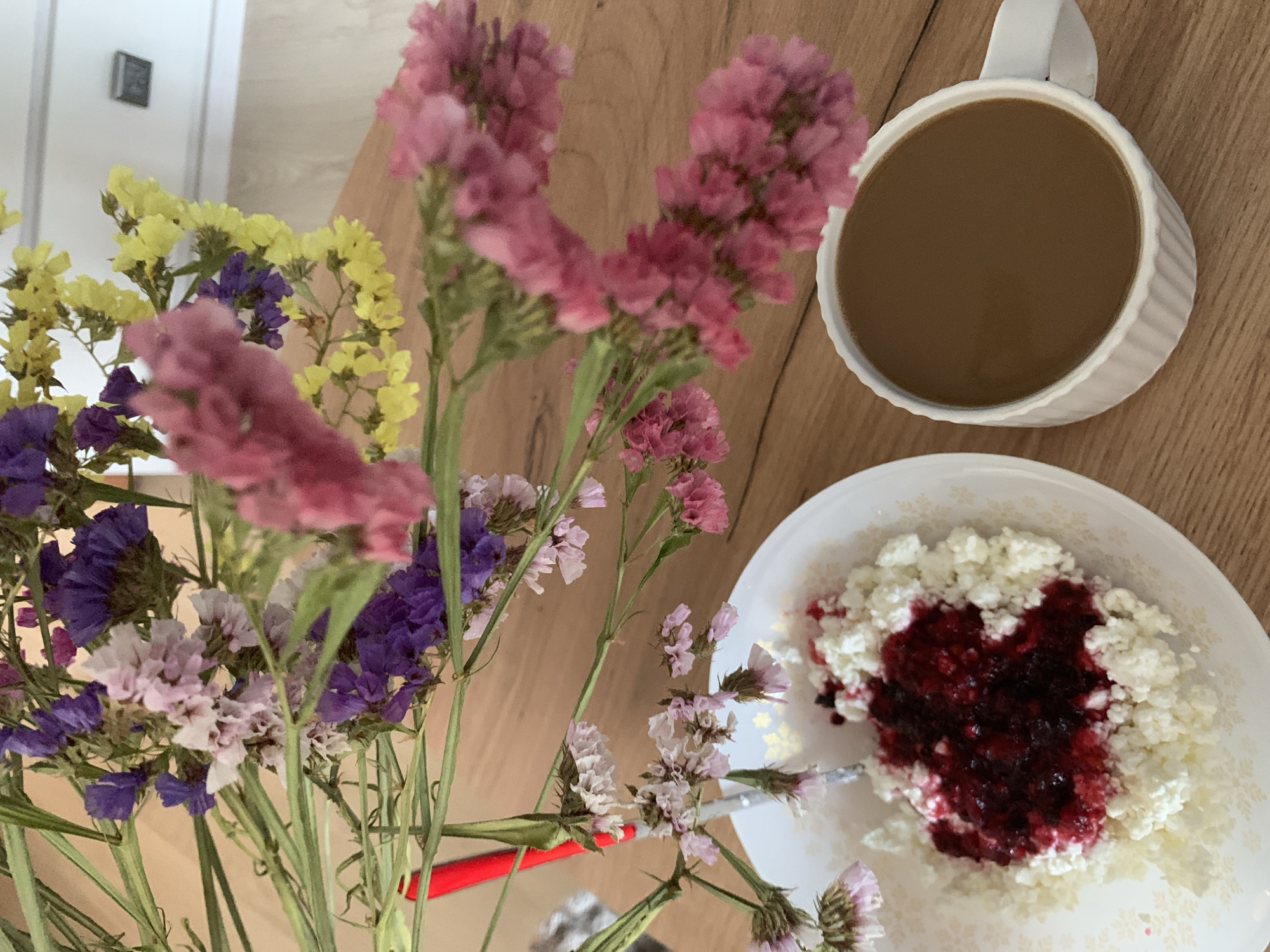 The image depicts a close-up, overhead view of a wooden table featuring a breakfast setting and a bouquet of dried flowers. In the lower left is a white bowl of cottage cheese topped with a dollop of dark red jam or compote. A silver spoon rests in the bowl. To the right of the bowl is a white ribbed mug filled with a light brown beverage, likely coffee. A bouquet of dried flowers, including purple, pink, and yellow blossoms, is arranged in the background, with some stems and flowers extending into the frame. The setting appears to be an indoor environment, potentially a home, with a blurred background suggesting a wall and possibly a light fixture. No people or animals are visible in the image. The scene is static and captures a moment of quiet breakfast. The image does not provide any information about the specific location within the city of Kapyĺ, Belarus.