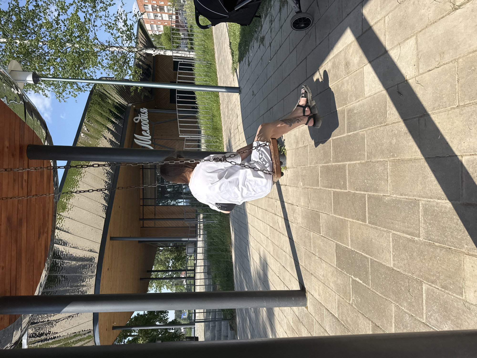 The image shows a person sitting on a swing in an outdoor area of what appears to be a park or recreational space. The person is wearing a white shirt and dark-colored sandals. They are seated with their legs extended, facing away from the camera. The swing is suspended by metal chains connected to dark grey metal poles.
The ground is paved with grey rectangular blocks. A stroller is partially visible in the background. In the distance, there is a building with the word "Melodiya" (Мелодия) visible on the façade in Cyrillic.
Given the mention of Votkinsk, Russia, it's likely the building is a cultural center, and the surrounding area is a public park or plaza, possibly near a cultural landmark. The setting appears to be sunny, based on the strong shadows cast on the pavement.