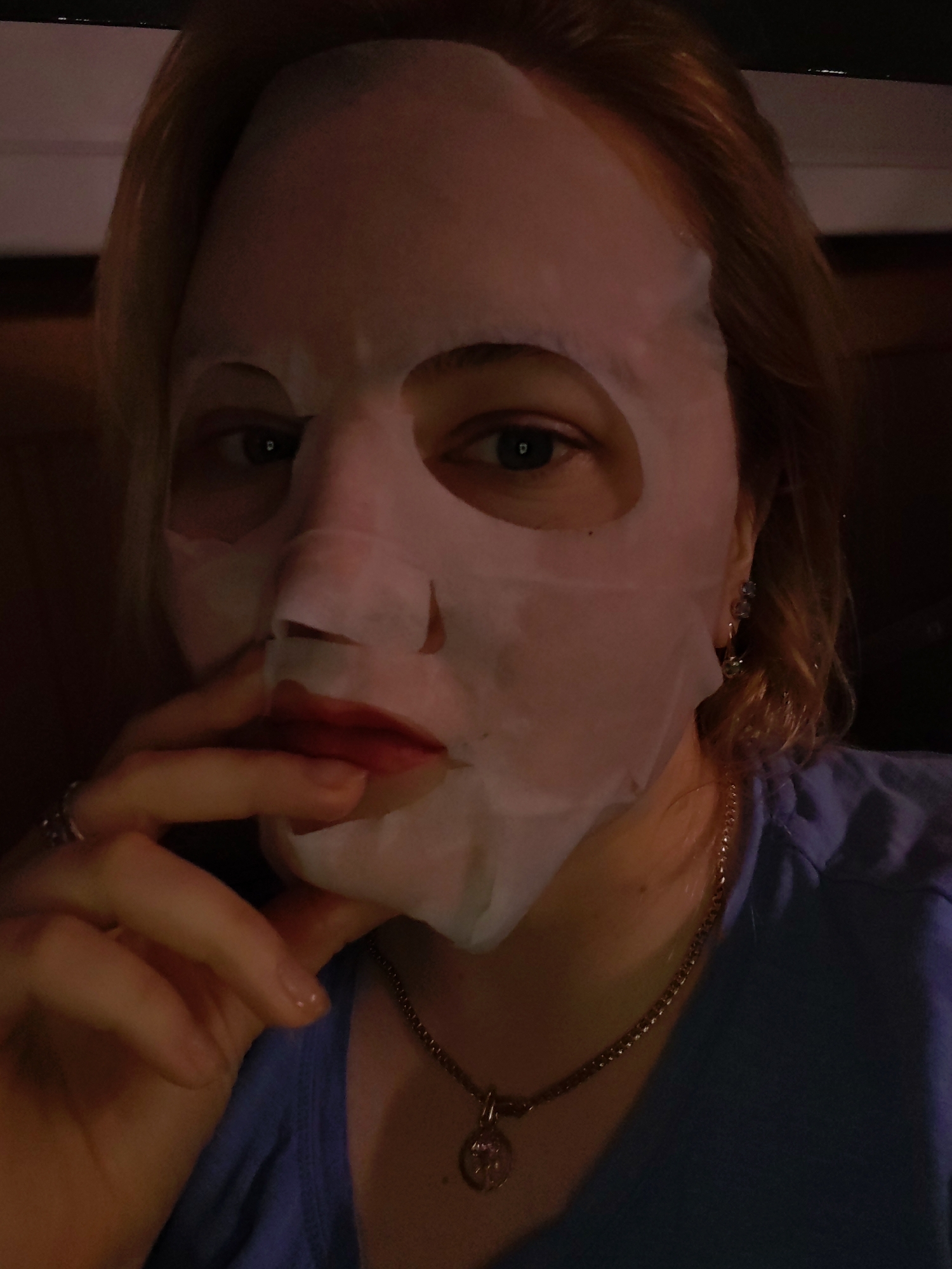 A woman with fair skin, wearing a blue v-neck shirt and a gold chain necklace with a round pendant, poses for a selfie while wearing a white sheet face mask. Her left hand is raised to her face, with her fingers touching her lips. She has light brown or blonde hair and is wearing earrings. The location is unspecified, and there is no evidence it is in the city of Izhevsk, Russia. The background is dark and indistinct.