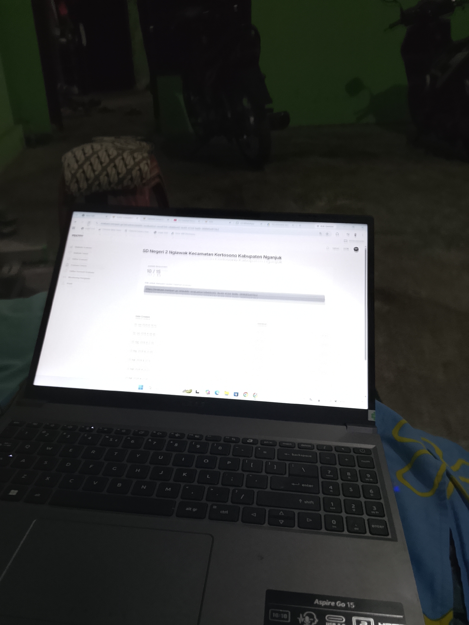The image shows a laptop screen displaying a webpage for "SD Negeri 2 Nglawak Kecamatan Kertosono Kabupaten Nganjuk." The webpage appears to be an online form or survey, with the text "10/15" visible, suggesting the user is on the tenth of fifteen items. Below this, there is a URL that seems to point to a survey or evaluation. Further down the page, there are timestamps indicating when the content was created or updated, with dates ranging from April 2023 to August 2023. The laptop is positioned on a surface, with a portion of a keyboard and trackpad visible in the foreground. In the background, the environment is dimly lit, with hints of furniture and what appear to be motorcycles. The overall setting suggests an indoor location, likely a home, in Kelurahan, Indonesia, based on the text on the screen.