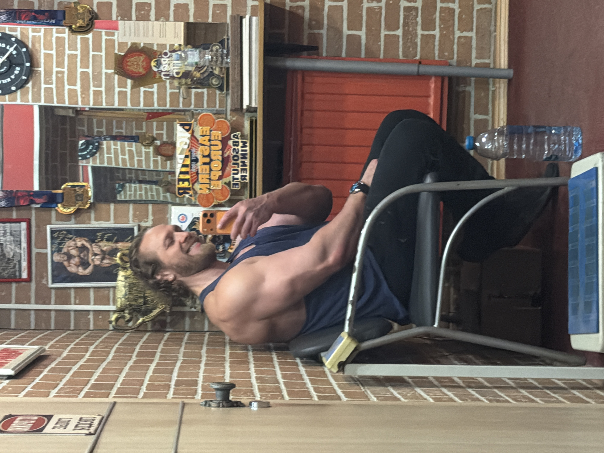 A muscular man with a beard and long hair is lying on a seated leg press machine in a gym. He is wearing a dark blue tank top and black pants. He is holding a smartphone in his hand and appears to be taking a selfie. A water bottle rests on the leg press machine. The gym has a brick wall decorated with fitness-related memorabilia, medals, posters, and a clock. A mirror is reflecting some of the wall decorations. There is a sign that appears to be written in Cyrillic. The location appears to be in Saint Petersburg, Russia, as implied by the Cyrillic signage and gym decorations that suggest a possible Eastern European theme. The man is likely taking a break or finishing his workout.