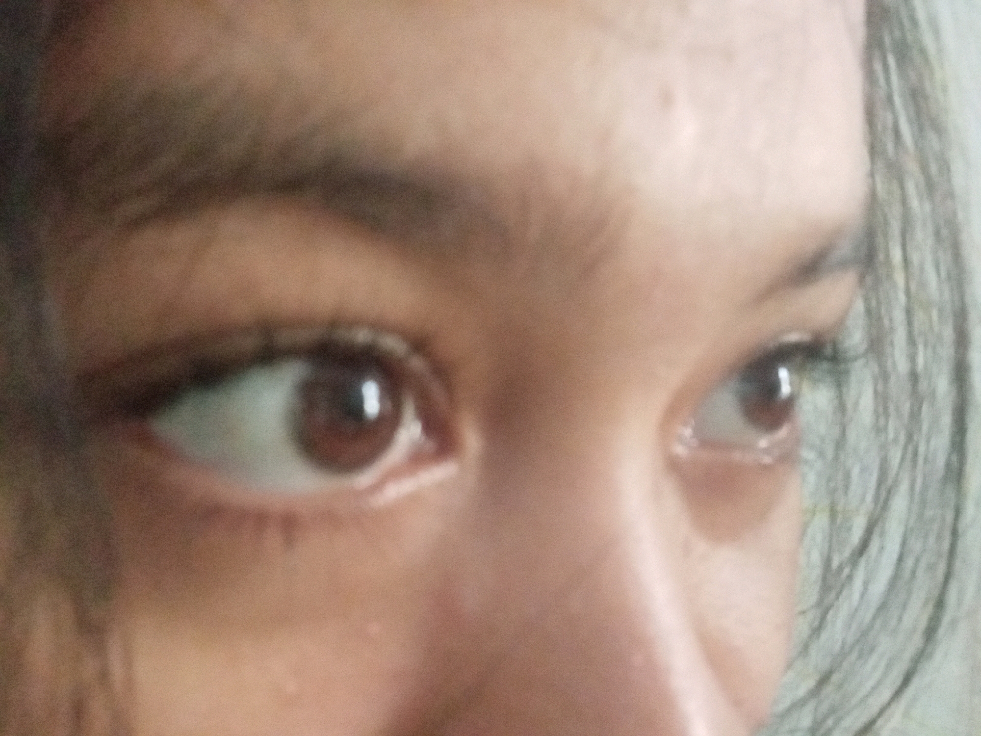 The image is a close-up, low-angle, and blurry photograph of the eyes of a young individual. The subject's face is partially visible, with the focus on their eyes. Their irises are a deep brown, and their eyebrows are dark and well-defined. The lighting appears to be natural, casting a soft glow on the skin. There are no discernible objects or environmental elements in the background, suggesting the photograph was taken indoors or in a controlled setting. The image does not contain any information that can be used to determine its location in Badas, Indonesia, or any other specific geographical area.