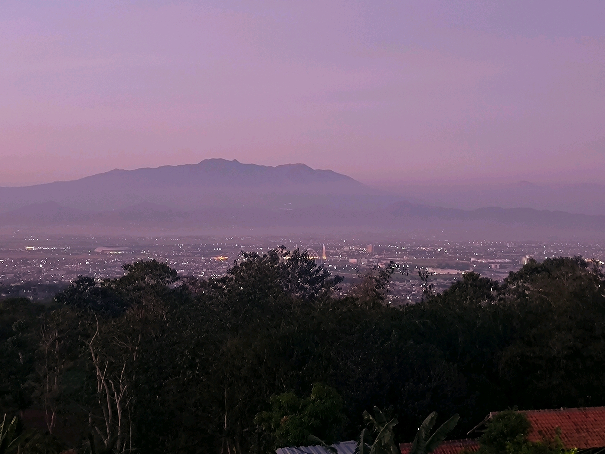 The image depicts a panoramic view of a city at twilight, possibly the city of Cilengkrang, Indonesia, as suggested by the provided context. In the foreground, a dense canopy of dark green trees partially obscures the view, with some tree branches and foliage visible. To the right, a portion of a reddish-tiled roof is present. Beyond the trees, the city lights twinkle across a vast expanse, creating a soft glow against the darkening landscape. In the distance, a mountain range is silhouetted against a gradient sky transitioning from a pale purple near the horizon to a deeper shade of purple and pink above. The overall atmosphere is serene and tranquil, captured during the transition from day to night. There are no people, animals, or any specific actions or interactions discernible in the image. The focus is on the natural landscape and the urban sprawl under a twilight sky.