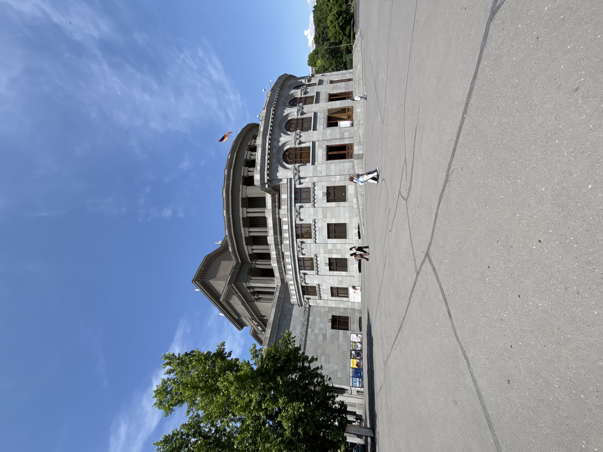 The image depicts the Yerevan Opera Theater, a prominent landmark in the city of Yerevan, Armenia. The building is a large, circular structure with classical architecture, characterized by its stone facade, arched windows, and a colonnaded upper level. A flag, likely the Armenian flag given its colors and placement on the building, is visible flying from the top of the structure. The building is situated in a wide, paved plaza, with distinct black lines marking sections of the pavement, suggesting a public square or event space.
Several individuals are present in the plaza, scattered across the open area. Two people are walking together in the foreground, and another person is walking in the middle ground to the right. A group of posters or advertisements are displayed on the exterior wall of the building on the left side, near a large tree with green foliage. The sky above is bright blue with sparse white clouds, indicating a clear, sunny day. The surrounding environment appears to be urban, with glimpses of trees and possibly other buildings in the distance to the right. The overall scene conveys a sense of public space and architectural grandeur under a daytime sky.