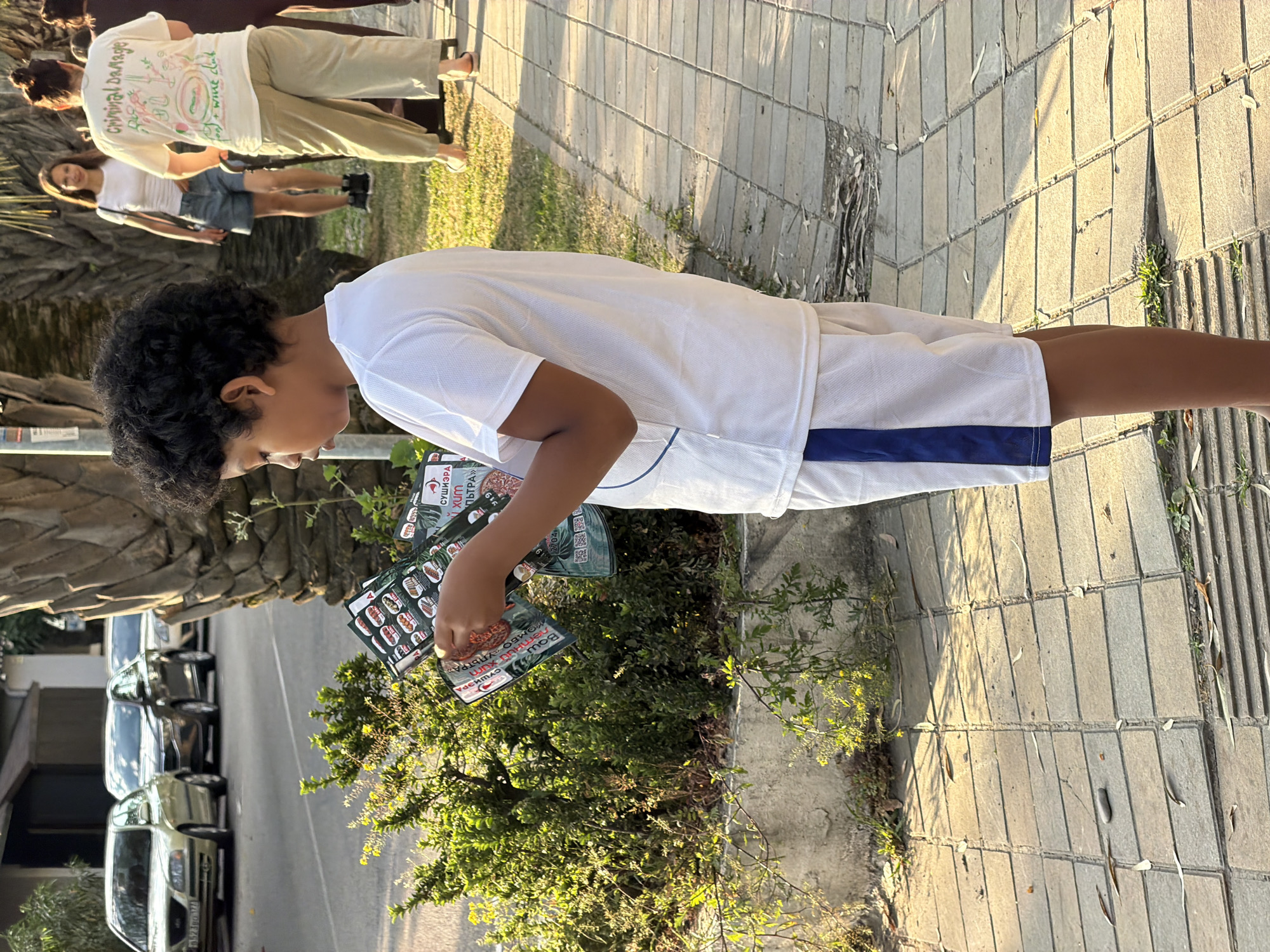 The image depicts a young boy of Afro-Caribbean descent standing on a paved walkway in Sochi, Russia. He is wearing a white t-shirt and white shorts with a blue stripe. The boy is holding a brochure, which appears to be a menu, open and is looking down at it. In the background, several cars are parked along a street. Further back, two women are walking on the sidewalk. One woman wears a white t-shirt with text and graphics, and the other wears a white t-shirt and denim shorts. The environment includes green bushes, palm trees, and a stone retaining wall. The ground is paved with rectangular stones. The scene is illuminated by natural light, suggesting it is daytime.