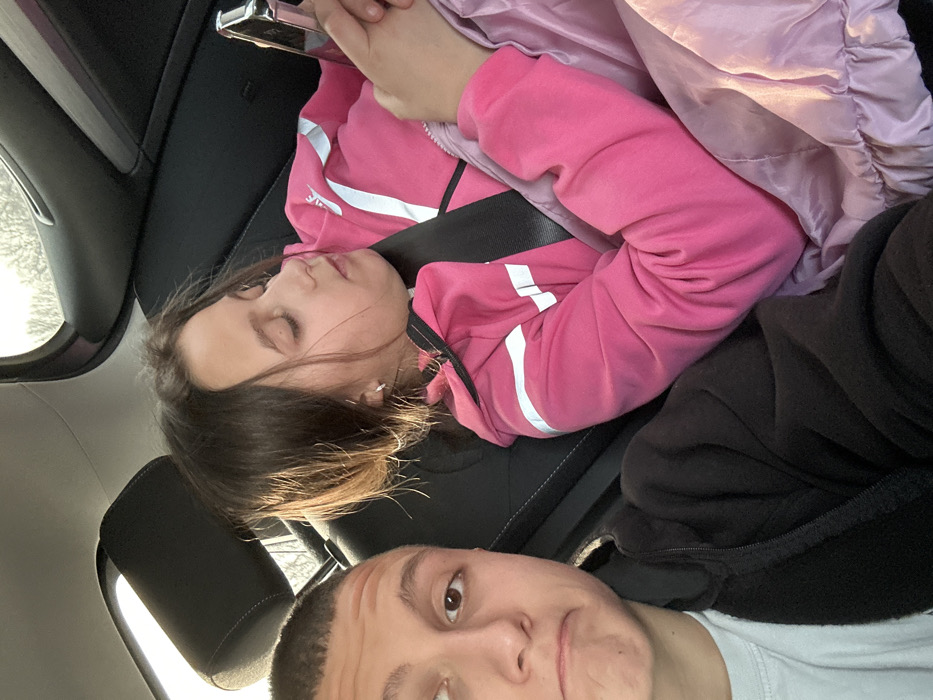 The scene is located inside the rear passenger compartment of a vehicle. The specific location within the city of Bolyarovo, Bulgaria, cannot be determined from the visual evidence.
Two individuals are present. On the left, partially visible, is a male with short, dark hair, light skin, and brown eyes. He wears a light-colored t-shirt under a dark, possibly black, zip-up fleece jacket. His head is turned slightly to the right, and his mouth is slightly downturned. On the right, a female with long, dark brown hair, light skin, and closed or nearly closed eyes, appears to be resting or engrossed in an object. She wears a bright pink hooded jacket with white reflective stripes on the arms, secured by a dark seatbelt across her chest and shoulder. She is also covered by a light purple or pink quilted jacket or blanket. Her hands are holding a silver-colored smartphone, with her thumbs positioned as if interacting with the device. Both individuals are seated in dark-colored car seats, visible are black headrests and portions of the car's interior trim. Sunlight is visible through the rear window, illuminating the female's hair and the car's interior.