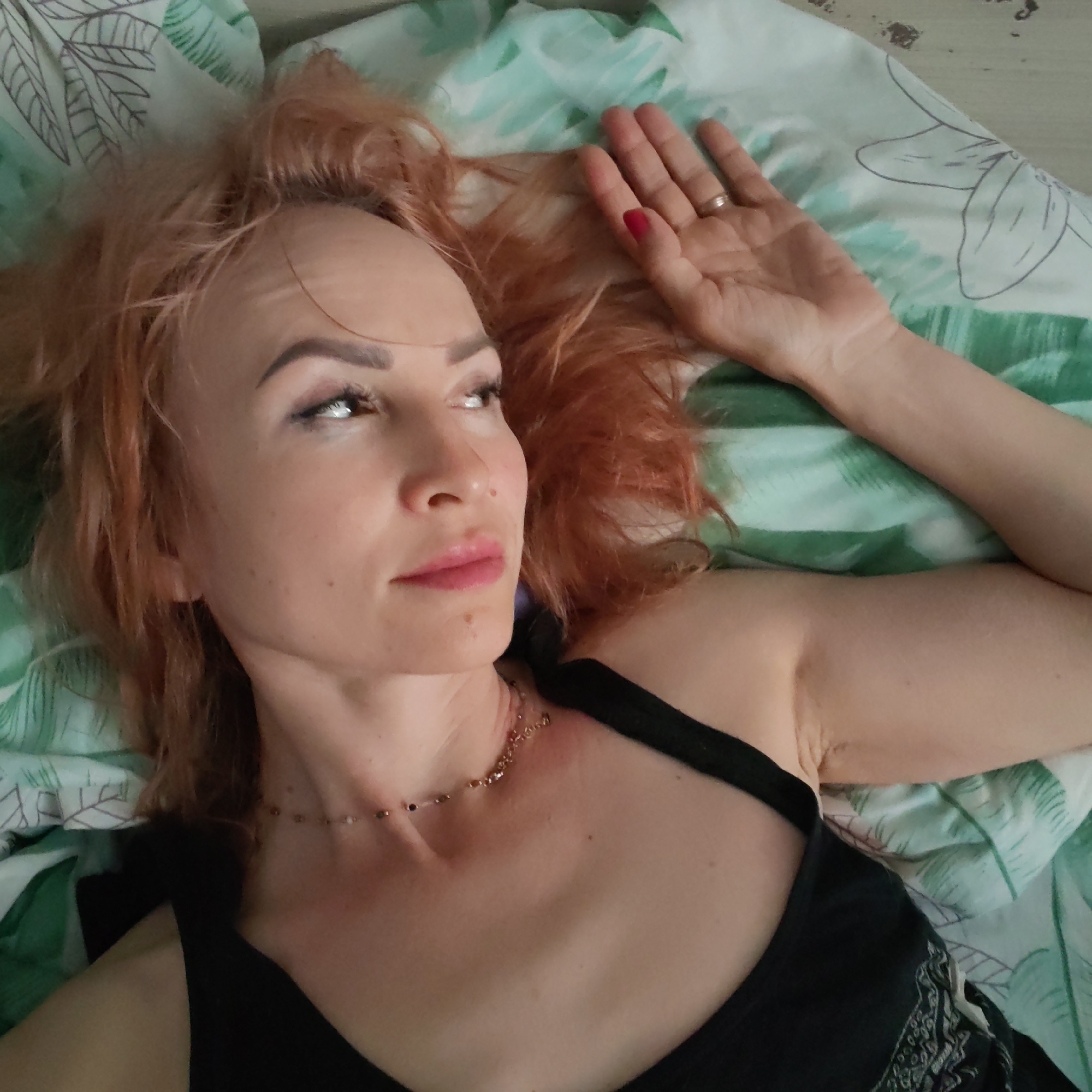 A medium-close-up shot shows a woman with reddish-blonde hair lying on a bed, likely in Chaikovsky, Russia. She is positioned diagonally across the frame, looking upwards and to her right with a thoughtful or pensive expression. Her light-colored hair is spread around her head on a green and white patterned pillow or duvet cover with tropical leaf motifs.
The woman has defined eyebrows, subtle eye makeup, and pink lipstick. A delicate, beaded necklace adorns her neck. She is wearing a black tank top or camisole, part of which shows a white or light-colored embroidered pattern near her right side. Her left arm is raised above her head, resting on the bedding, revealing a simple metallic ring on her left ring finger and red nail polish on her fingernails. Her skin appears fair, with a few small moles visible on her face and neck. The overall lighting is soft, suggesting an indoor setting. There is no visible text in the image.