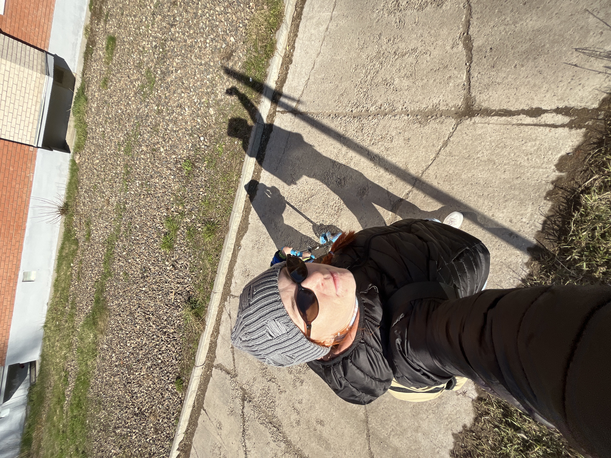 A first-person, top-down perspective photo shows a person standing on a concrete sidewalk in Chaikovsky, Russia. The person is wearing a gray knit cap, sunglasses, and a black puffer jacket. The shadow of the person, including the camera and arm extended to take the photo, is cast on the sidewalk. Adjacent to the sidewalk is an area covered in small rocks and sparse grass. In the background, there is a building with brickwork on the exterior. The setting appears to be a sunny day.