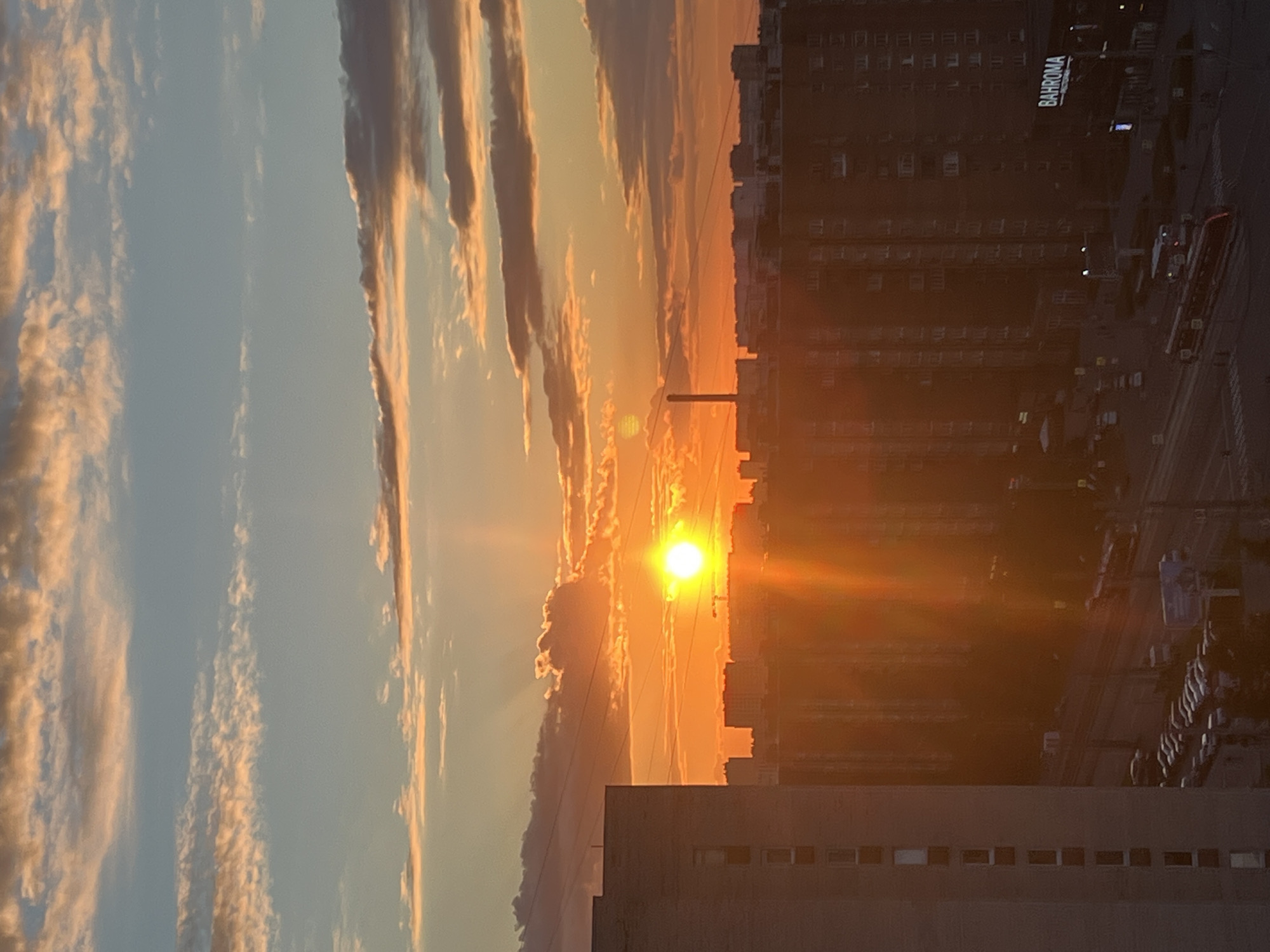 This is a photograph of a sunset over a city in Russia. The sun is low in the sky, casting a warm orange glow over the buildings and clouds. The clouds are a mix of pink, orange, and blue, with some wispy white clouds scattered throughout. The city below is a silhouette of buildings, with some streetlights visible. A tram is seen on the street. The overall impression is one of a peaceful and beautiful end to the day.