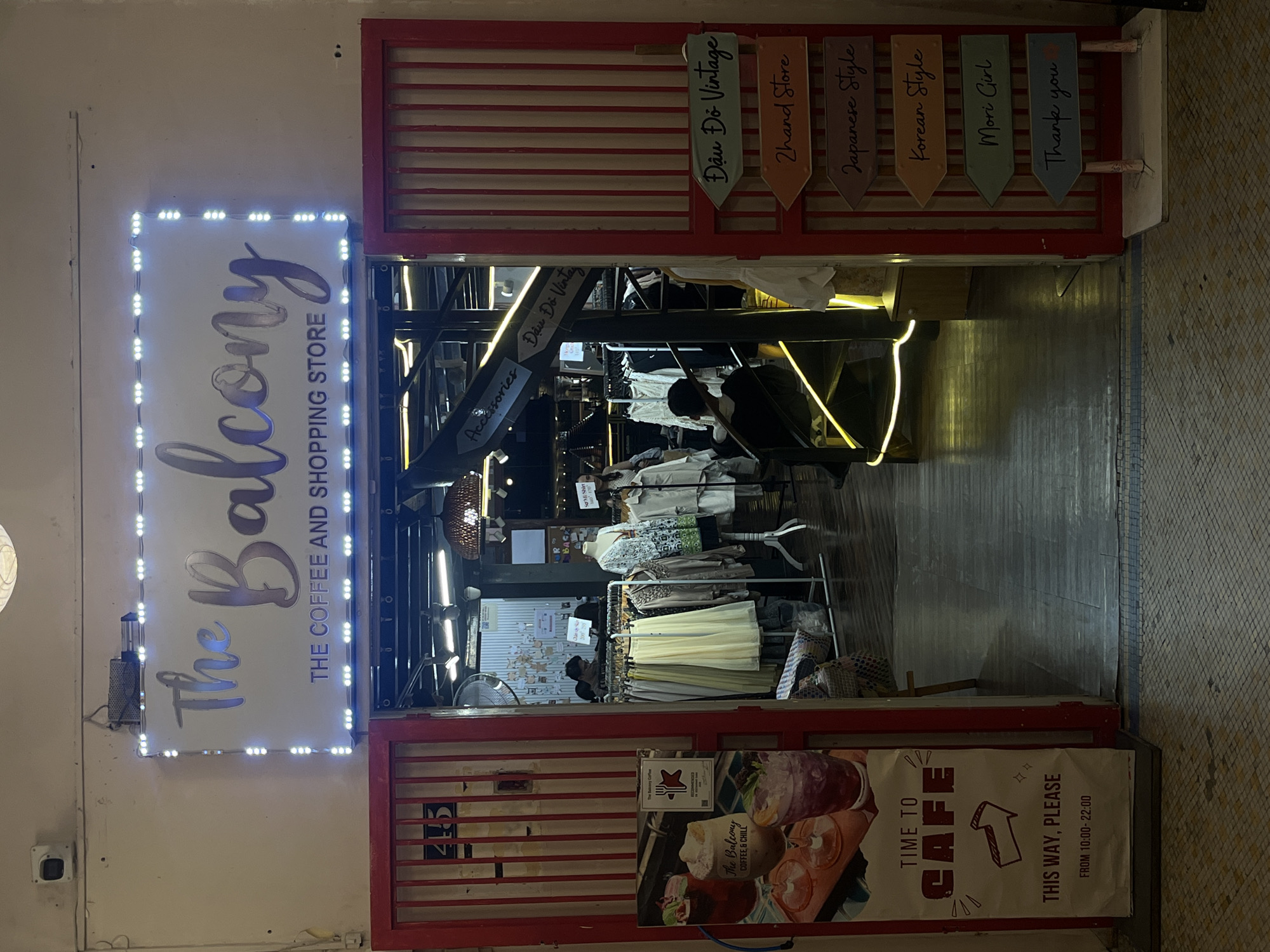 The image shows the entrance to "The Balcony," a coffee and shopping store located in Thủ Đức, Vietnam. The storefront features a prominent sign with the store's name in stylized lettering, illuminated by white LED lights. Below the name, the text "THE COFFEE AND SHOPPING STORE" is visible. Red-barred doors flank the entrance.
To the left of the entrance, a vertical poster displays images of drinks and text including "TIME TO CAFE" and "THIS WAY, PLEASE," along with operating hours from 10:00 to 22:00.
Inside the store, clothing racks are visible, displaying various garments and mannequins. A spiral staircase with yellow accent lighting is also present. Wooden directional signs are mounted to the right of the entrance, indicating categories such as "Đâu Đỏ Vintage," "2hand Store," "Japanese Style," "Korean Style," "Mori Girl," and "Thank you."
The overall environment suggests a retail establishment with a focus on fashion and beverages, likely in a commercial area.