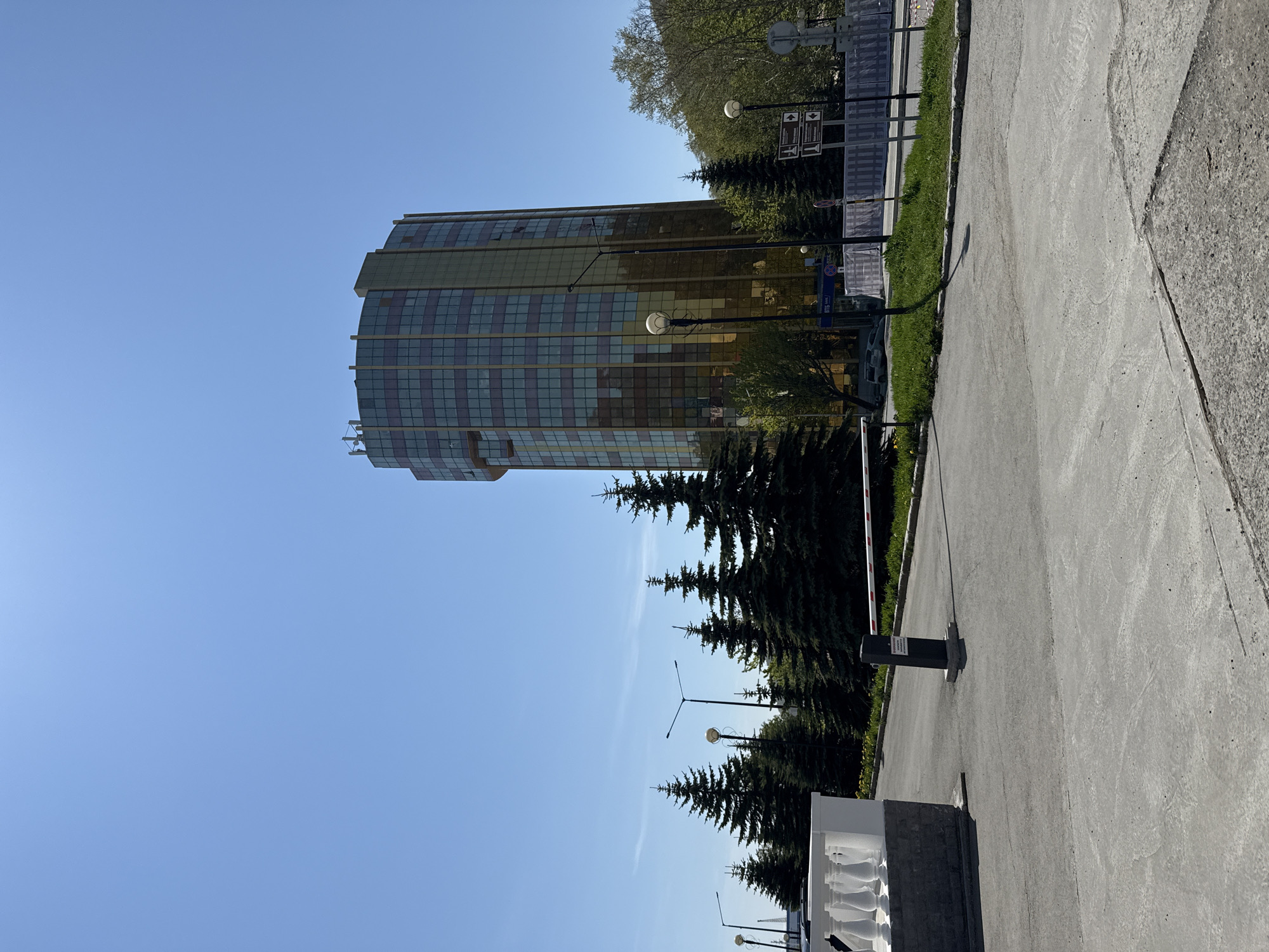 In Petropavlovsk-Kamchatsky, Russia, a modern, cylindrical building with gold-tinted windows stands tall against a clear blue sky. The building features a distinctive facade with a grid-like pattern of windows. In the foreground, a paved area leads towards the building, bordered by a row of tall, dark green evergreen trees. A white balustrade is visible to the left. A white and red barrier arm, typically used for access control, is lowered across the paved area. Several streetlights are positioned along the road. The environment appears to be an urban setting with a mix of greenery and modern architecture.