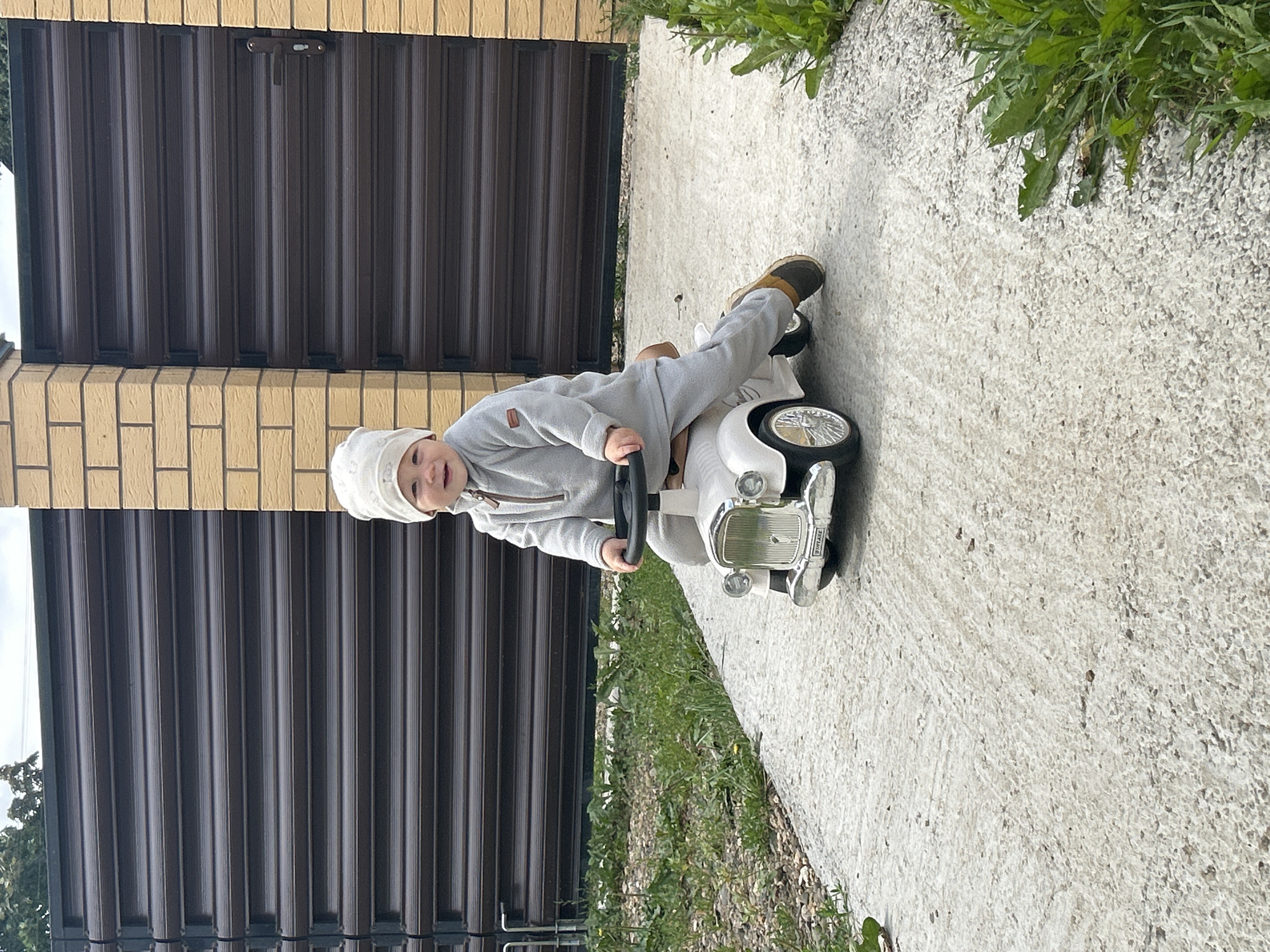 A young child, wearing a light-colored beanie and a grey matching tracksuit, sits on a white toy car with chrome accents and a black steering wheel. The child is smiling and looking towards the camera. The toy car is on a light grey concrete path. To the left of the path is a patch of green grass with some pebbles. To the right of the path, more green grass and weeds are visible. In the background, there is a dark brown metal fence with horizontal slats, supported by a brick pillar on the right side of the image. The scene appears to be outdoors, possibly in a residential area or yard. The location is identified as Chaikovsky, Russia, suggesting a typical urban or suburban setting within that city.